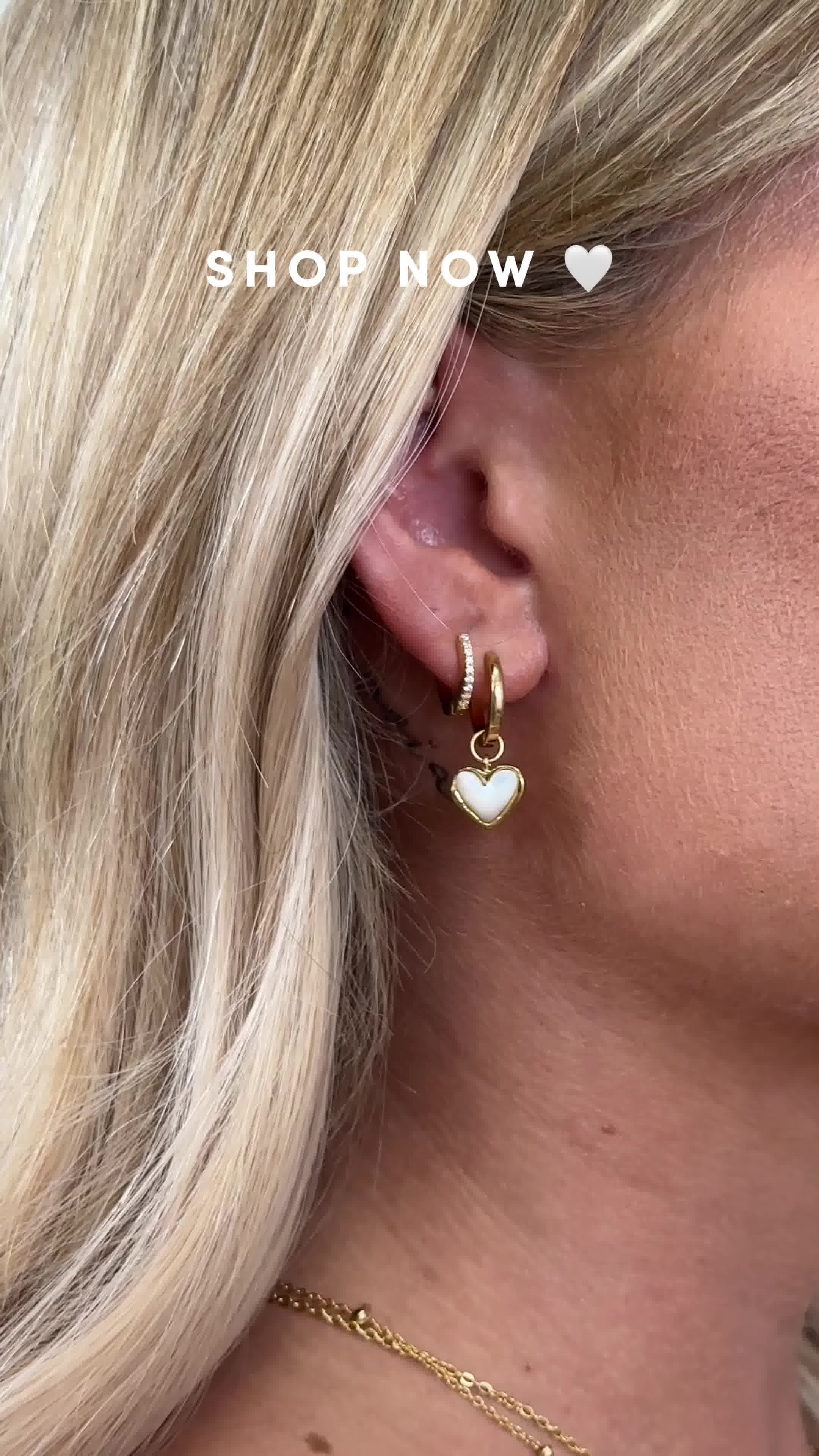 A close-up shot focuses on a woman's ear adorned with delicate gold hoop earrings, one featuring a small white heart-shaped charm. The background is softly blurred, highlighting the jewelry. White text overlaid on the image reads "SHOP NOW" with a small heart icon next to it, encouraging immediate purchase.