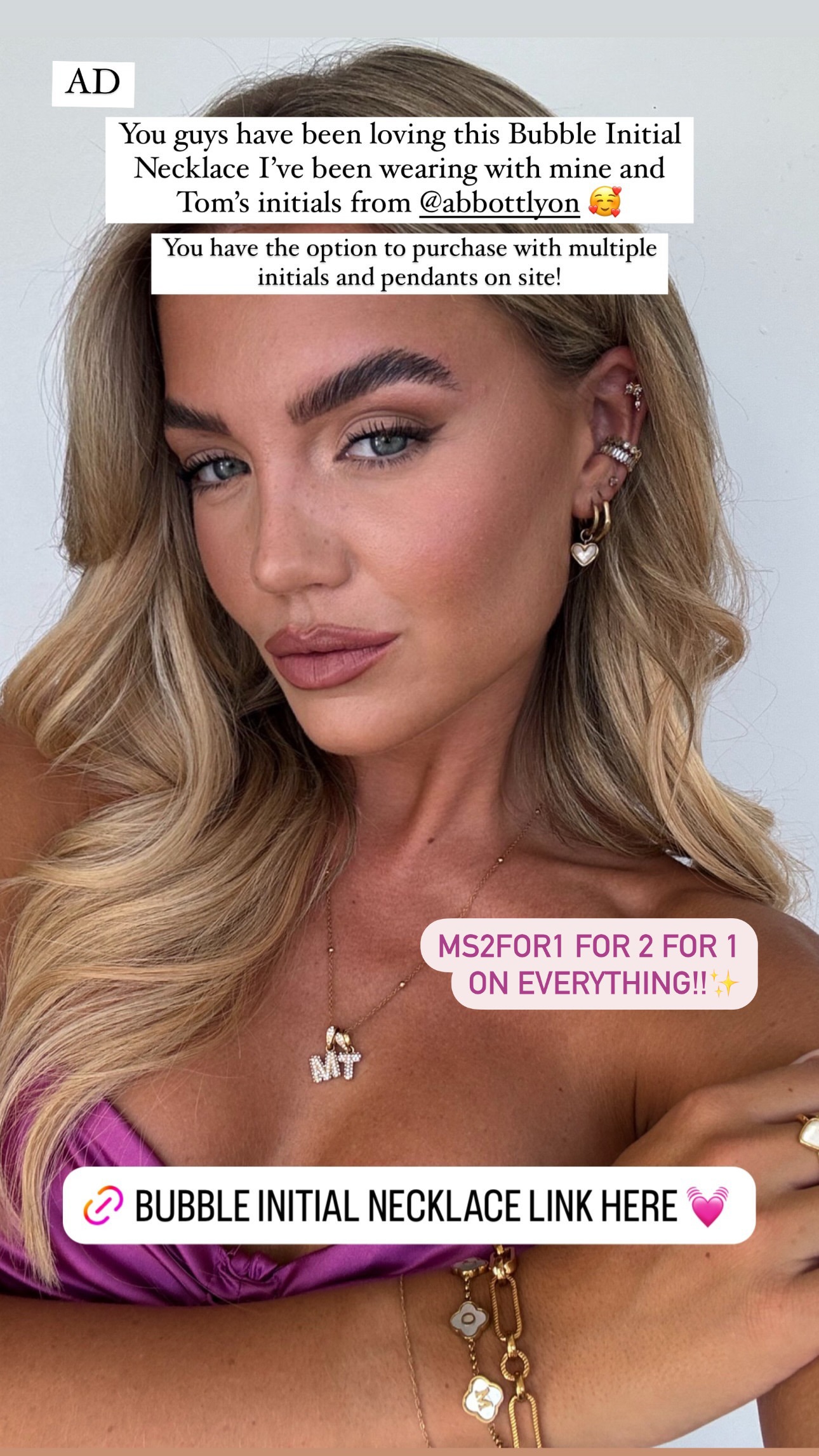 A close-up, shoulder-up shot of a woman with blonde wavy hair and full makeup. She is wearing a gold necklace with two initial charms, 'M' and 'T'. Text overlays on the image read 'AD', 'You guys have been loving this Bubble Initial Necklace I've been wearing with mine and Tom's initials from @abbottlyon 🥰', 'You have the option to purchase with multiple initials and pendants on site!', and 'MS2FOR1 FOR 2 FOR 1 ON EVERYTHING!! ✨'. Another text overlay at the bottom reads 'BUBBLE INITIAL NECKLACE LINK HERE 💕'.