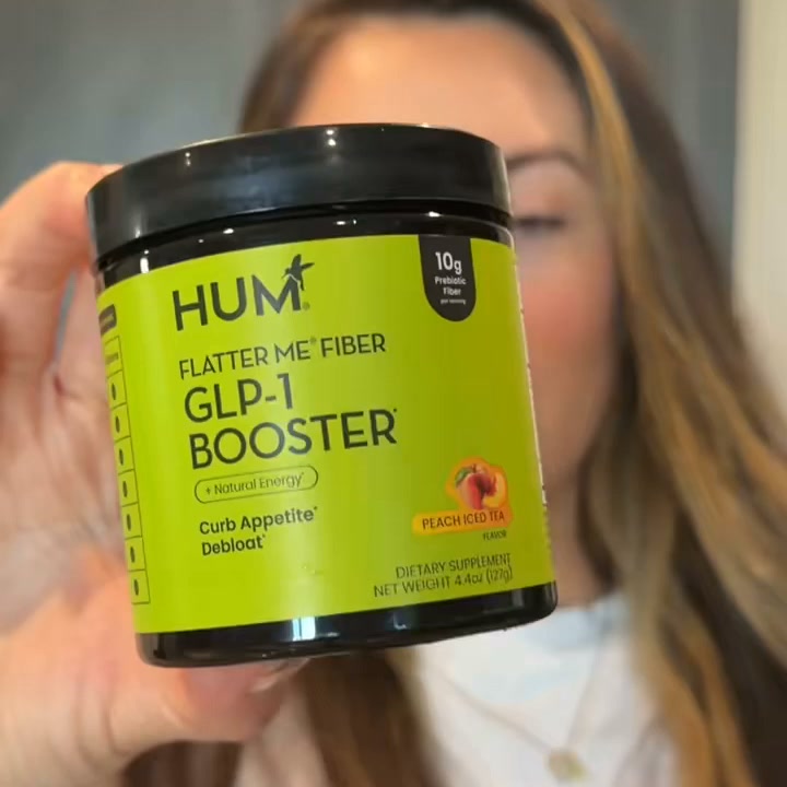 A person's hand holds up a jar of HUM Flatter Me Fiber GLP-1 Booster. The jar is bright green with black text. On-screen text highlights "10g Prebiotic Fiber" and "+ Natural Energy". The flavor is "Peach Iced Tea". The background is blurred, showing part of a person's face and hair. The creative focuses on the product's nutritional information and benefits.