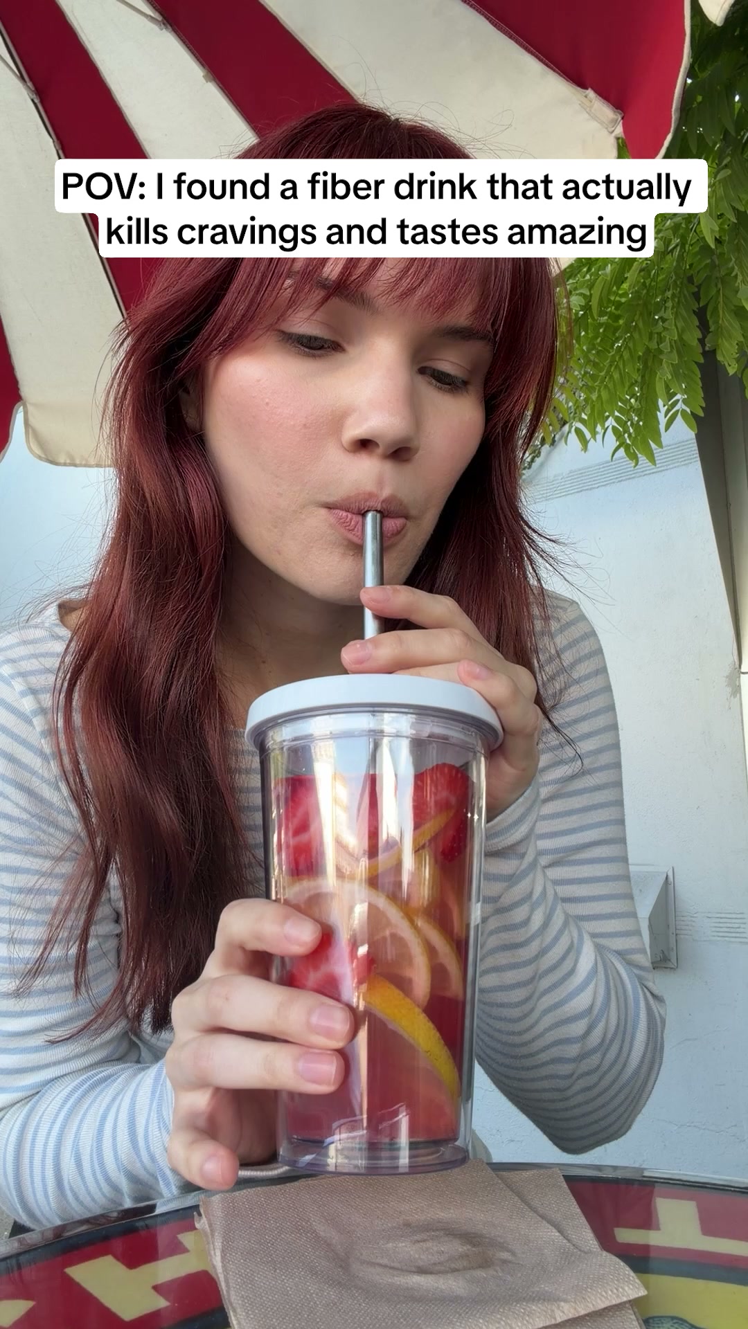 A young woman with reddish-brown hair is shown from the chest up, drinking from a clear tumbler filled with water, strawberries, and lemon slices. She is wearing a striped long-sleeved shirt. White text overlaid on the top left of the image reads: "POV: I found a fiber drink that actually kills cravings and tastes amazing." The overall scene suggests a casual, outdoor setting, possibly a cafe, with a hint of greenery and a striped umbrella in the background.