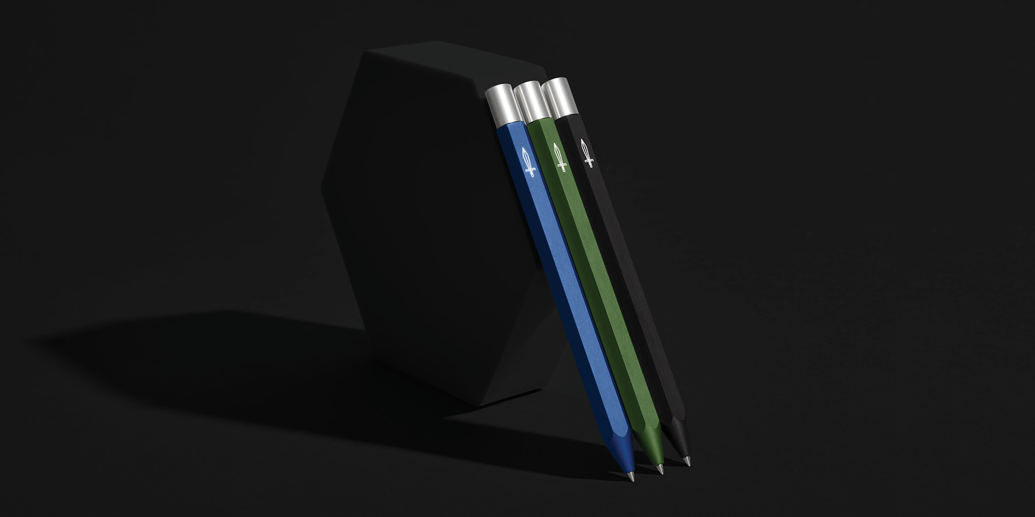Three sleek, hexagonal pens in blue, green, and black are arranged in a row, leaning against a dark, geometric object. The pens have silver caps and a small, white sword emblem near the top. The background is a dark, minimalist studio setting with dramatic shadows. The text overlay highlights the pen as Baronfig's best yet, built on customer feedback and legacy.