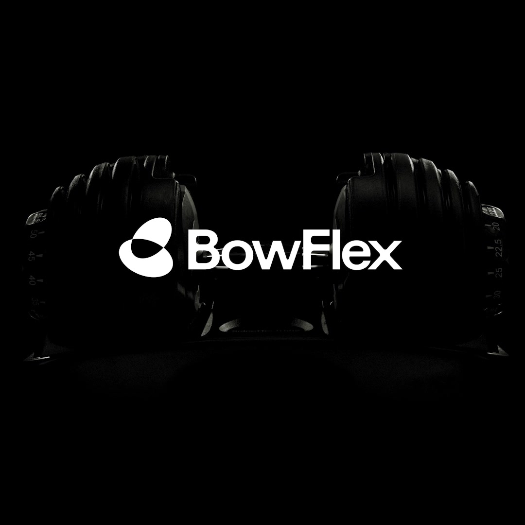 The ad features a dark, moody shot of two Bowflex adjustable dumbbells, positioned symmetrically with the Bowflex logo prominently displayed in white text between them. The lighting highlights the contours of the dumbbells, emphasizing their design. The text overlay, which also serves as the hook, describes the innovative and adaptive nature of the equipment, suggesting a personalized and intuitive workout experience. The overall aesthetic is sleek and modern, focusing on the product itself.