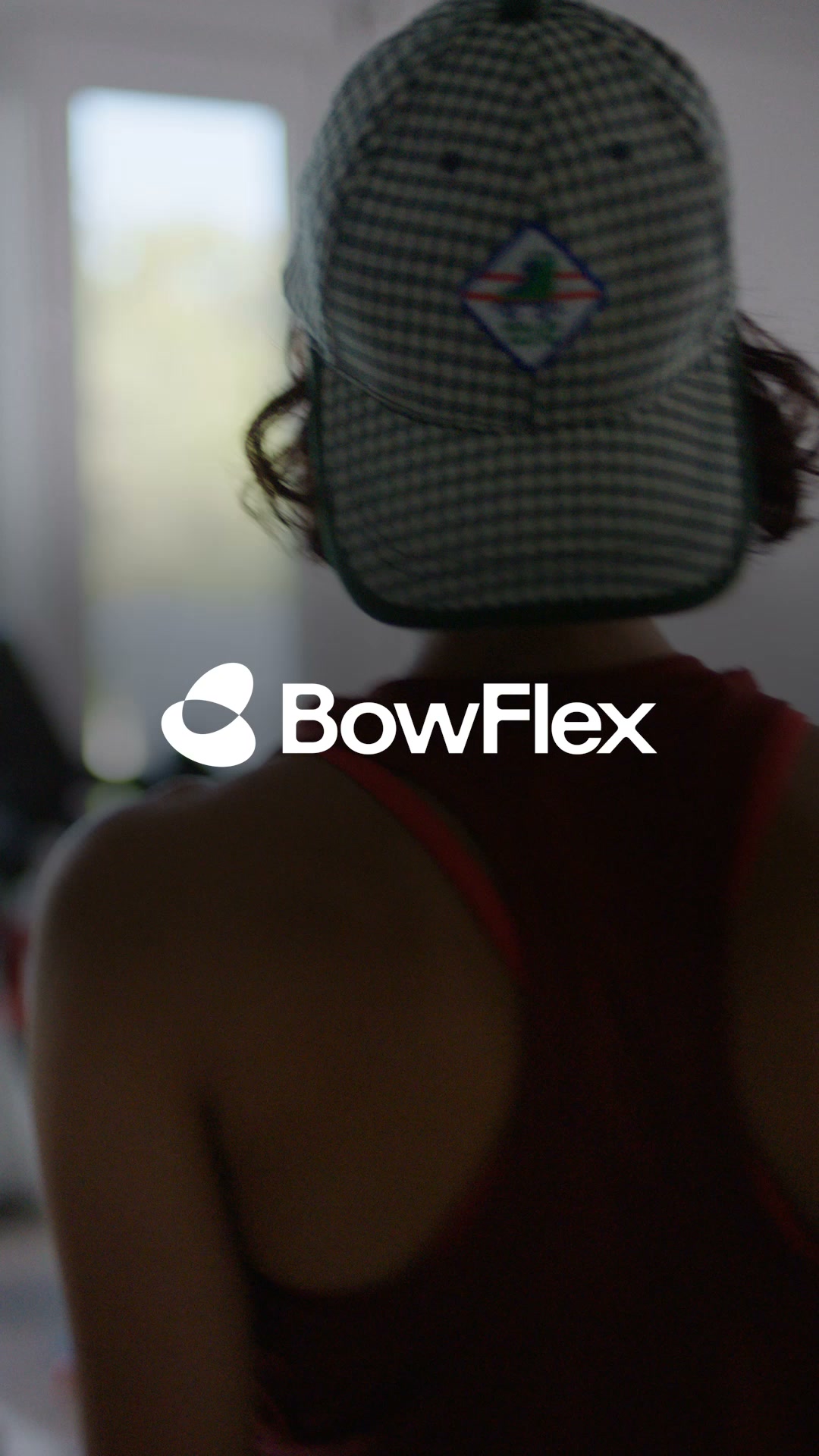 The creative features a close-up, from behind, of a person wearing a checkered baseball cap and a maroon tank top. The BowFlex logo is prominently displayed on the right side of the image. The text "Find your strength with BowFlex." is overlaid on the image, with a muscle emoji following the text. The background is blurred, suggesting an indoor setting, possibly a gym.