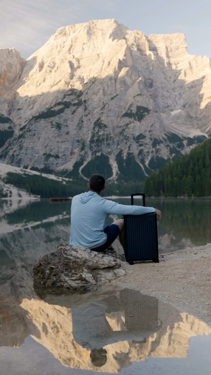A man sits on a rock by a calm lake, facing away from the camera, with a large, rugged mountain range in the background. He is wearing a light blue hoodie and dark shorts, with a dark-colored Nomatic suitcase beside him. The scene is reflected in the water, creating a symmetrical view. The text overlay reads: "#nomaticpartner It’s time to experience luggage you want to love, not leave."