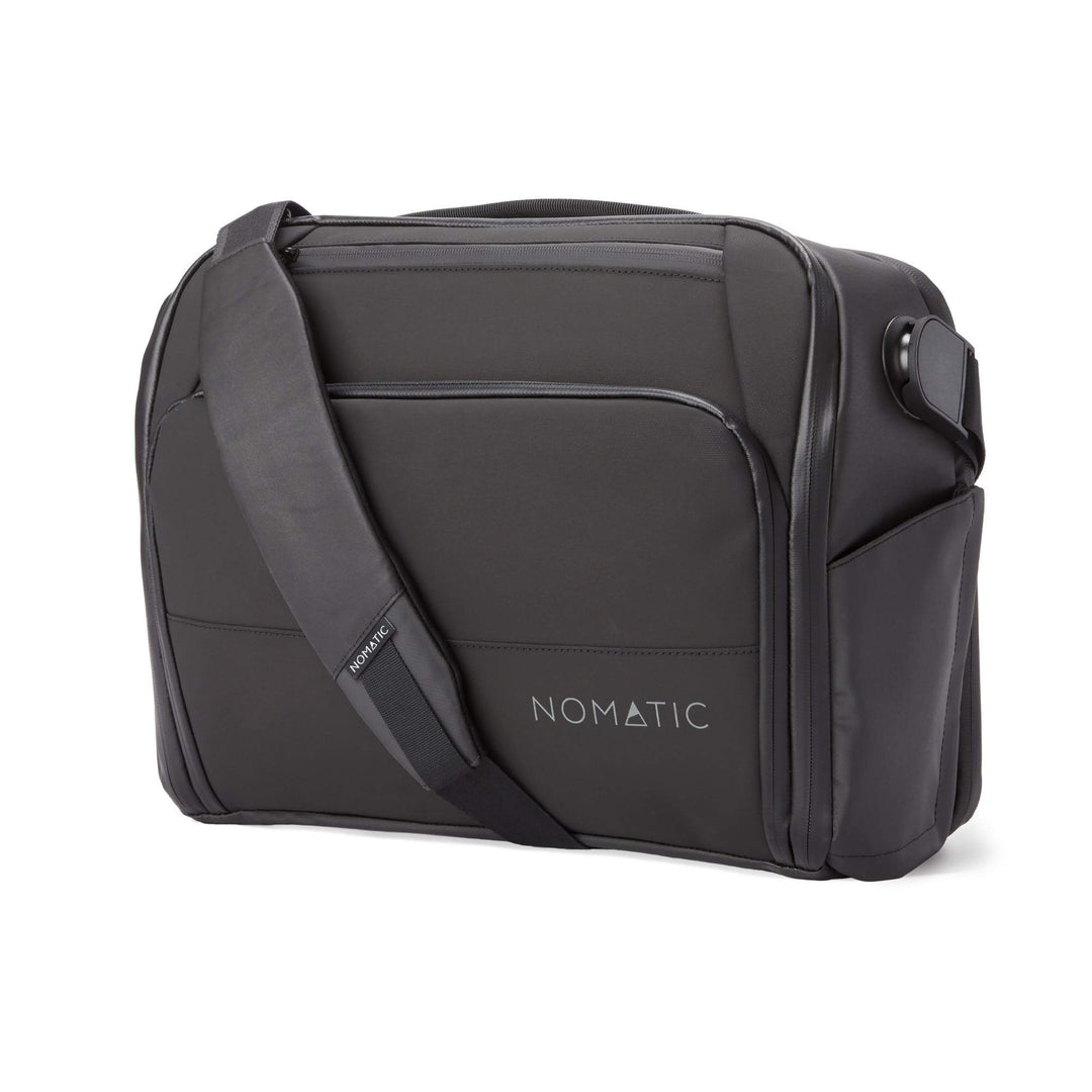 A black Nomatic messenger bag is shown from a three-quarter angle against a white background. The bag has a prominent shoulder strap with the Nomatic logo visible. The Nomatic logo is also subtly embossed on the front of the bag. The image is clean and focuses solely on the product's design and branding, with no additional text or graphics overlayed on the image itself.