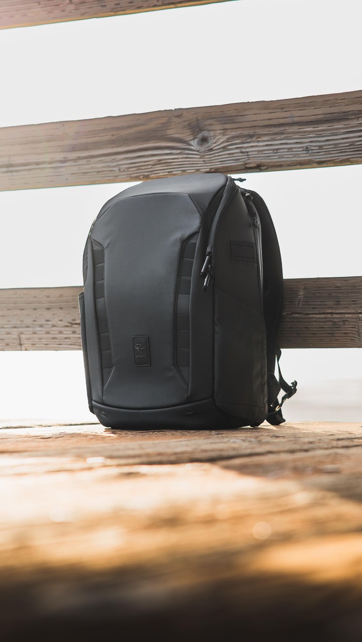 A black Nomatic 25L camera backpack is displayed outdoors on a wooden surface, with a blurred background of water and sky. The backpack is the primary focus, showcasing its textured material and zippers. Overlay text at the top reads "The Gamechanger ☝️", and the copy mentions an unboxing of the product, highlighting its storage and customization features.