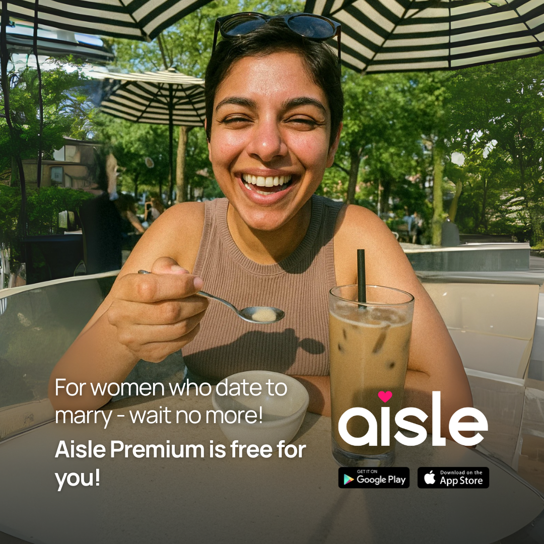 A woman with short dark hair and a wide smile is shown from the chest up, holding a spoon with a small amount of food and looking directly at the camera. She is seated outdoors at a table with a glass of iced coffee and a small white bowl in front of her. The background shows trees and umbrellas. Overlay text reads: "For women who date to marry - wait no more! Aisle Premium is free for you!" The Aisle logo and app store download buttons are also visible.