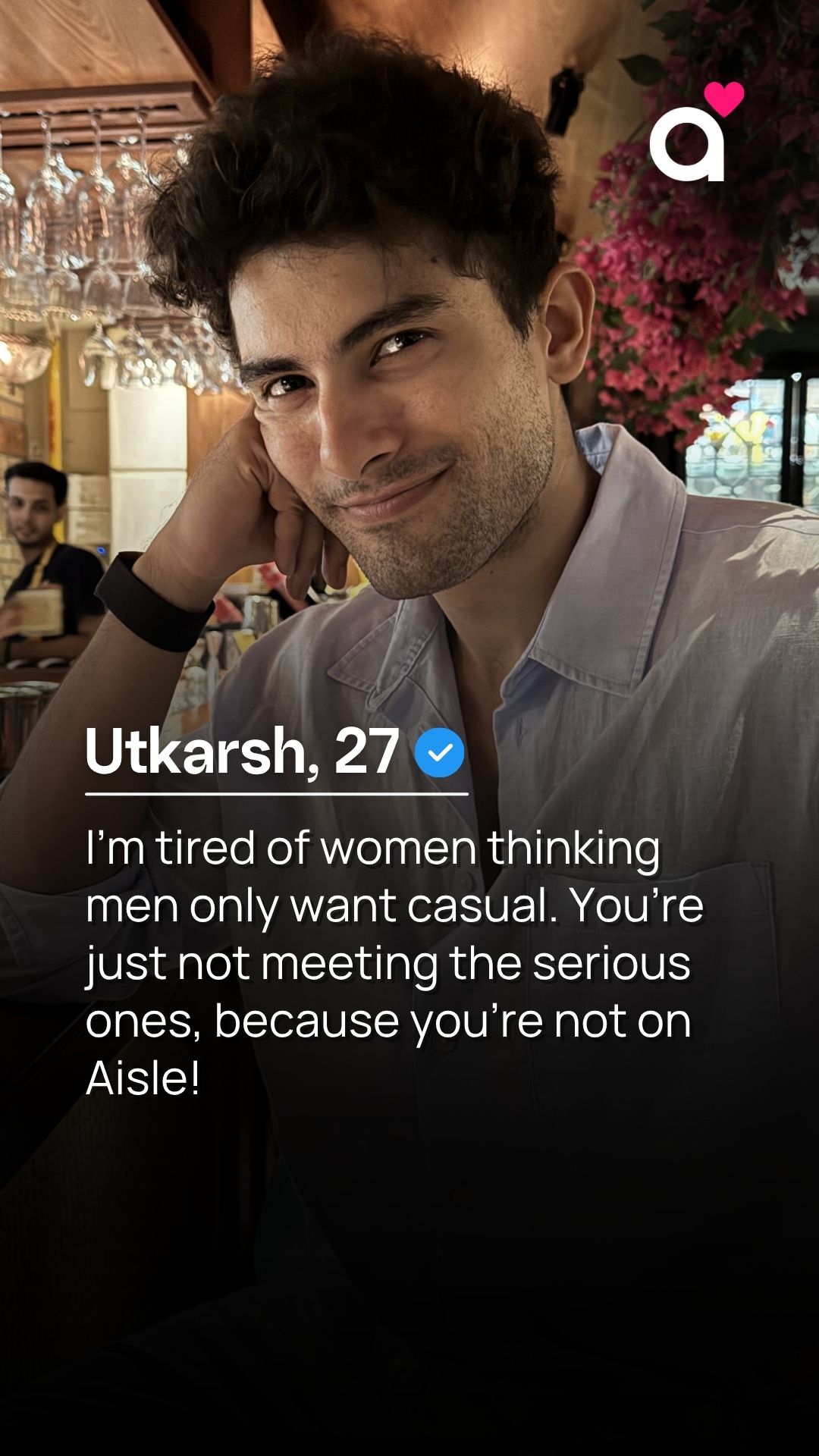 A man named Utkarsh, aged 27, is shown leaning his head on his hand and looking at the camera with a slight smile. He is wearing a light blue collared shirt. Text overlays his image, stating his name and age, followed by a statement about relationships and a mention of the Aisle app. The background appears to be a dimly lit bar or restaurant with hanging glasses and pink flowers.