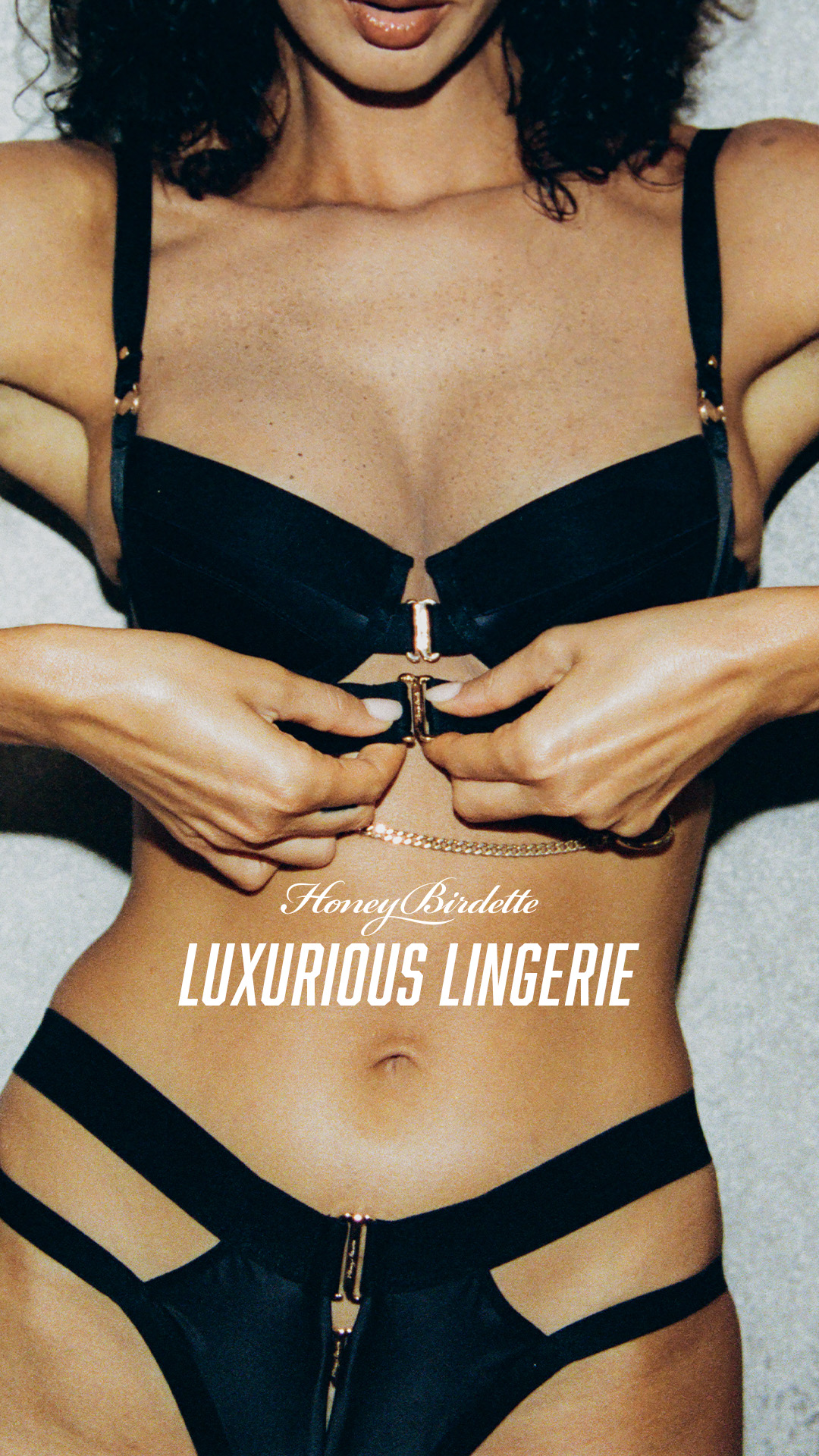 A close-up shot of a woman wearing black lingerie, with her hands fastening the bra clasp. White text overlays the image, reading "Honey Birdette" in a script font and "LUXURIOUS LINGERIE" in a bold sans-serif font. The focus is on the intricate details of the lingerie and the model's body, emphasizing luxury and sensuality.