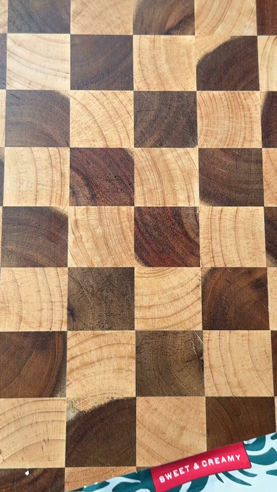 A close-up, top-down view of a wooden cutting board with a checkerboard pattern of light and dark wood squares. A red and white label with the text "SWEET & CREAMY" is visible in the bottom right corner, partially overlapping the wood. The image is a product shot with text overlay, highlighting the product's name and implying a desirable taste profile.