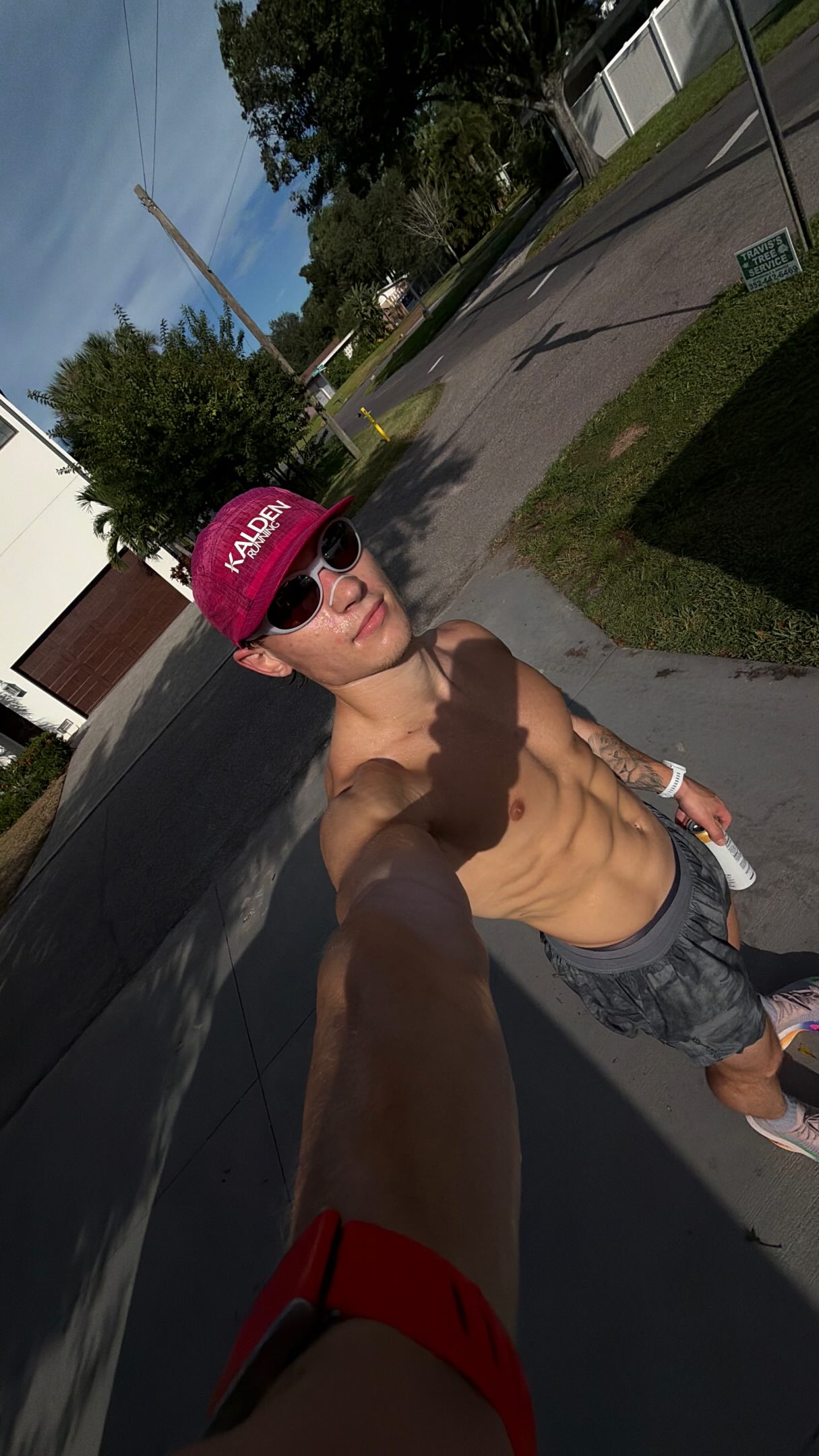 A young man, appearing to be a runner, is filming himself in a selfie-style video outdoors on a sunny day. He is shirtless, wearing a pink 'KALDEN RUNNING' hat, sunglasses, and athletic shorts. He is holding a can of what appears to be a beverage. The background shows a suburban street with houses, trees, and a sidewalk. Text overlays on the video state 'Hydration isn’t optional. It’s the foundation of how the whole day goes, miles included.' and 'One scoop of @lairdsuperfood Hydrate Coconut Water and I’m out the door. Hydration the way nature intended. 🥥 #ad #LairdSuperFoodPartner'.