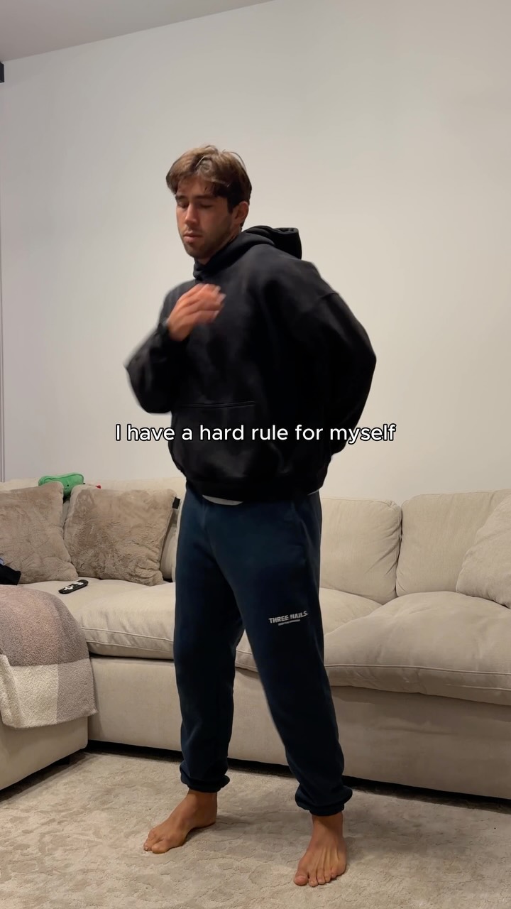 A man in a black hoodie and dark sweatpants stands in a living room, looking down and to his left with his hand on his chest. White text overlaid on the image reads "I have a hard rule for myself". The background shows a couch and a wall. The man is barefoot on a rug.