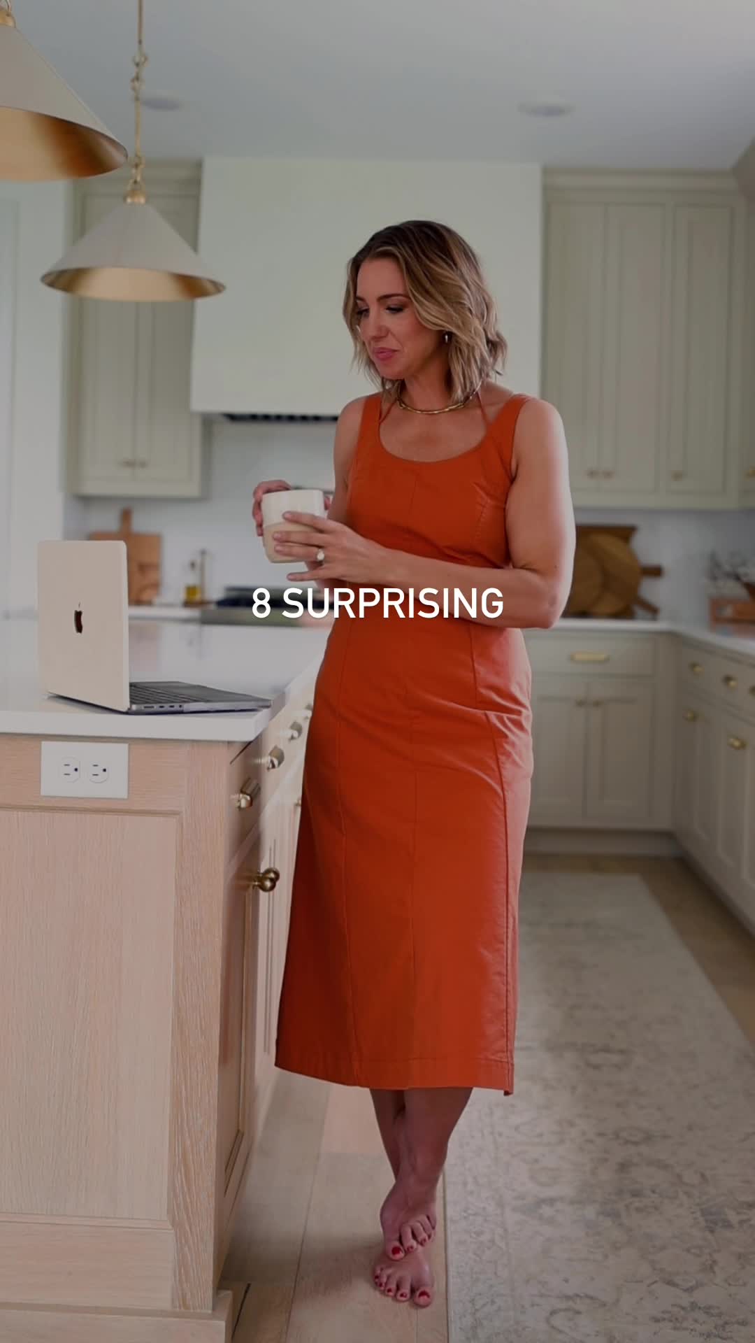 A woman in an orange dress stands in a modern kitchen, holding a mug and looking at a laptop. White text overlaid on the image reads "8 SURPRISING". The background shows kitchen cabinets and a stove hood. The overall scene is bright and airy, suggesting a professional or home office environment.