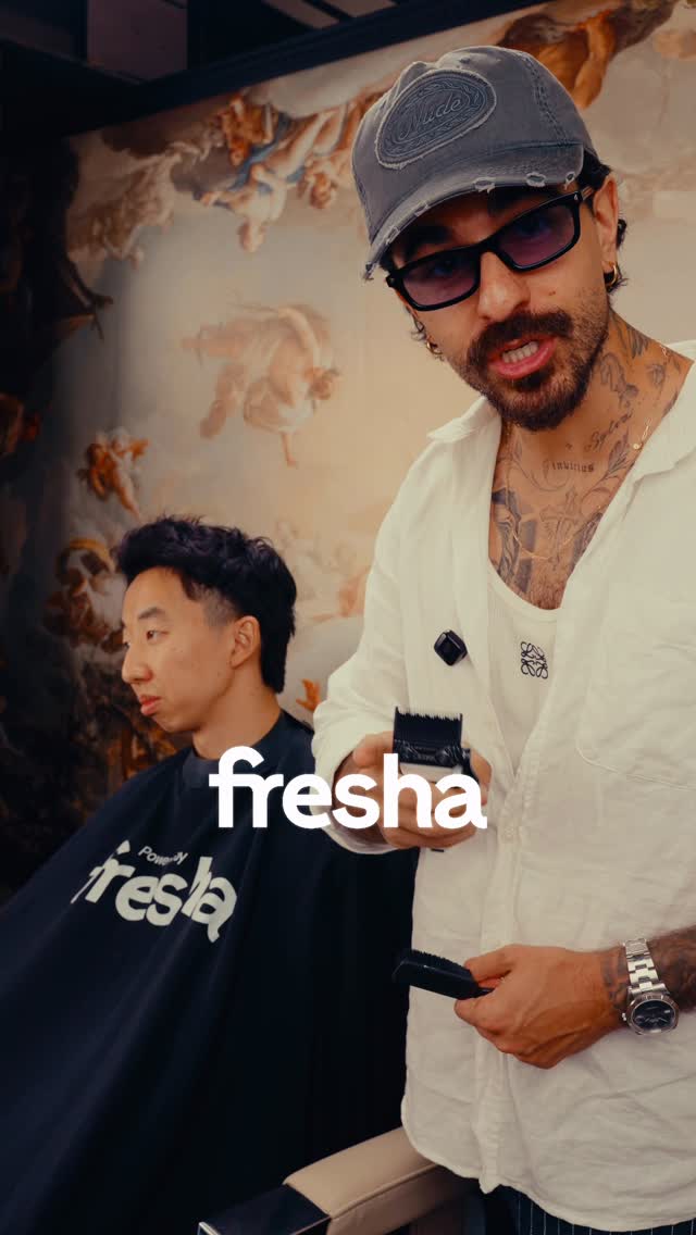 A barber wearing a distressed baseball cap and sunglasses is holding clippers and a comb, looking at the camera. He is standing in front of a client who is seated in a barber chair, wearing a black cape with the Fresha logo. The background features a classical mural. White text overlay reads "fresha" across the center of the image.