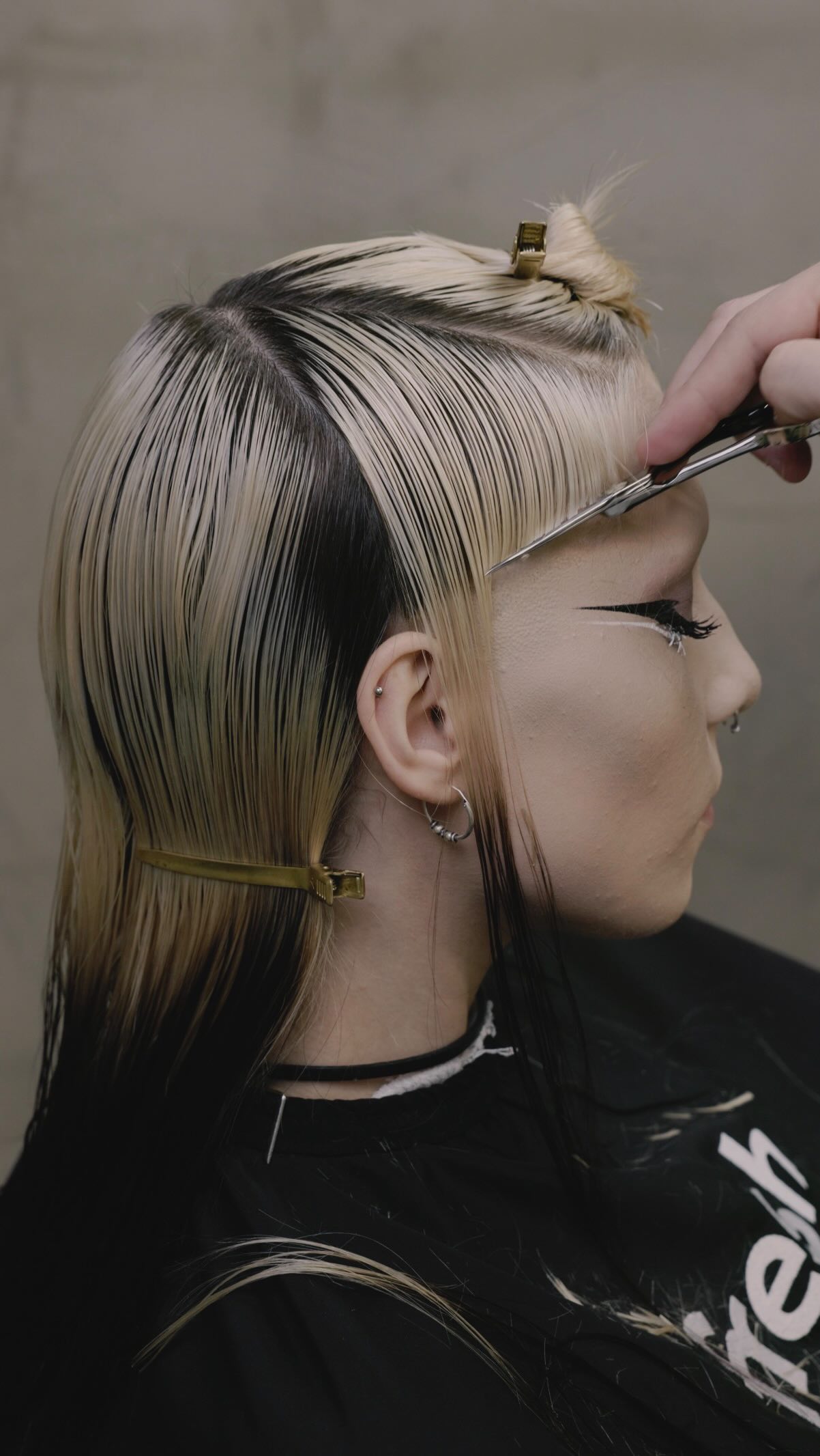 A close-up profile view of a person with dyed blonde and black hair getting a haircut. A hand is holding scissors and trimming the front section of the hair. The person has dramatic winged eyeliner and nose piercings. The background is a plain, textured wall. The text on the creative reads: "12k likes on the previous post. As promised this is the step by step. Sometimes the most beautiful things come randomly, but when you work with a system you know you can’t fail."
