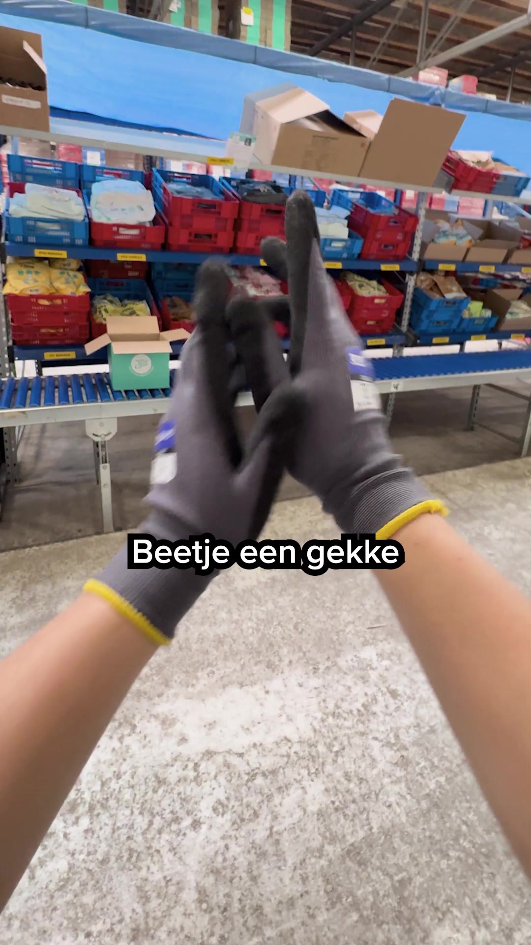 The video shows a first-person perspective of someone wearing grey work gloves, clapping their hands together in front of a warehouse setting with shelves stocked with red and blue crates. White text overlayed on the bottom of the screen reads "Beetje een gekke". The scene suggests a behind-the-scenes look at a fulfillment or distribution center.