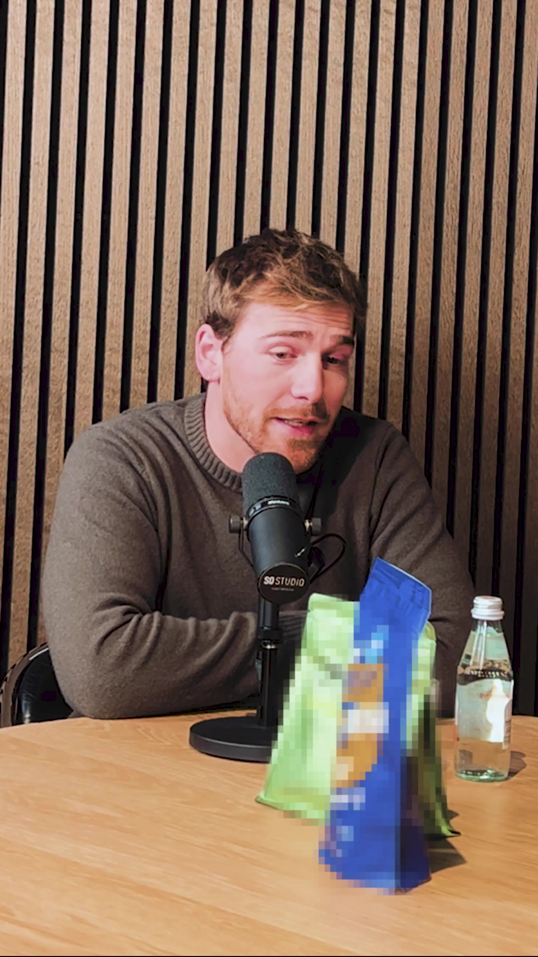 A man with short, light brown hair and a beard is sitting at a wooden table, speaking into a microphone. He is wearing a dark grey sweater. Behind him is a wall with vertical wooden slats. On the table in front of him are two pixelated bags, one green and one blue, and a clear bottle of water. The text on the screen says "30 seconden voor een perfect ontbijt! ⚡️ Ontdek hoe je in no-time een eiwitrijke maaltijd met weinig suiker maakt! Voor maar €1,59 p/maaltijd" followed by bullet points detailing nutritional information and a risk-free trial offer.