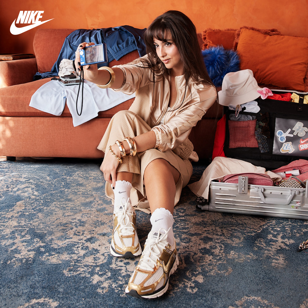 A young woman is sitting on the floor, taking a selfie with a digital camera. She is wearing a beige satin tracksuit and white socks with Nike sneakers. In front of her is an open suitcase filled with clothes and accessories, and a couch with clothes draped over it. The Nike logo is visible in the top left corner of the image. The text "Add a retro touch to any fit." is overlaid on the image.