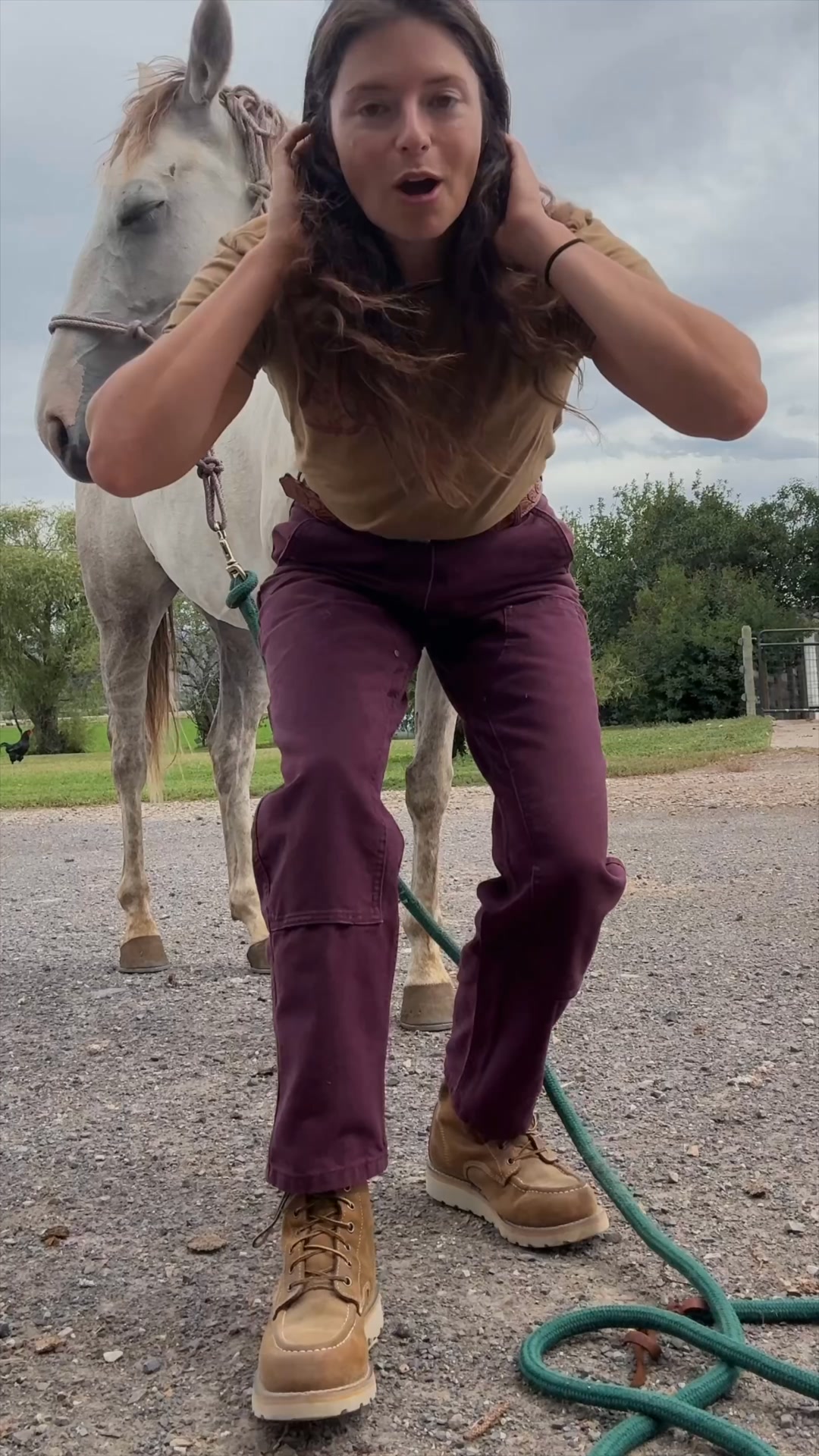 A woman with long, dark, wavy hair stands next to a white horse. She is wearing a tan t-shirt and dark purple pants, with tan work boots. Her hands are on her head, and her mouth is open in a surprised or excited expression. The background shows a gravel path, green grass, trees, and a cloudy sky. The text overlay reads: "Farm chores in the morning, standing on concrete all day, and still presentable enough for a meeting."