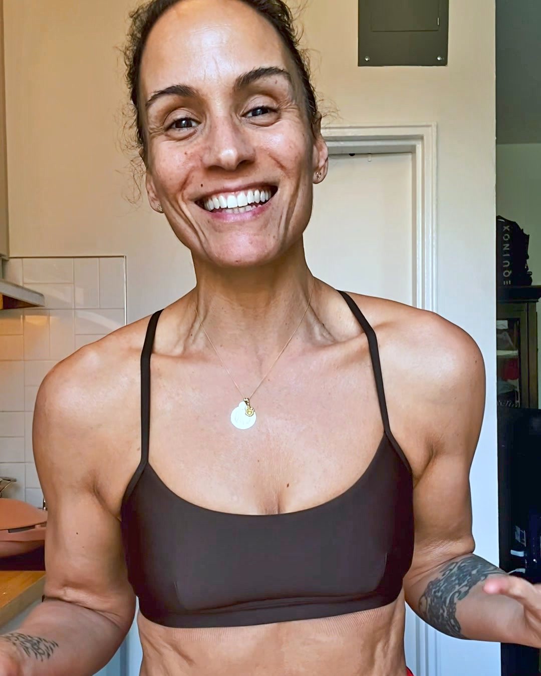 A woman with a beaming smile and athletic build is shown from the chest up, wearing a dark sports bra and a delicate necklace. She is looking directly at the camera with a confident and energetic expression. The background is a softly lit indoor setting, possibly a kitchen or home gym. Text overlays on the image ask the viewer to consider their aging process and introduce 'Basis' as a solution for cellular health.