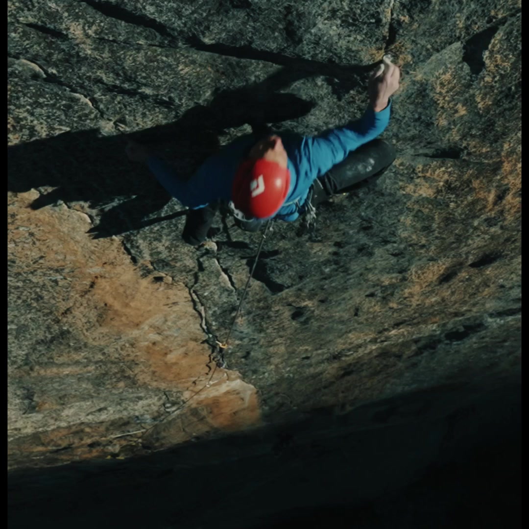 A climber in a red helmet and blue jacket ascends a steep rock face. The camera is positioned directly above, looking down at the climber. Sunlight casts a dramatic shadow on the rock. Text overlays the image, stating "Between you and the summit: the right equipment. Equipment for alpinists." The scene emphasizes the challenging nature of climbing and the importance of gear.