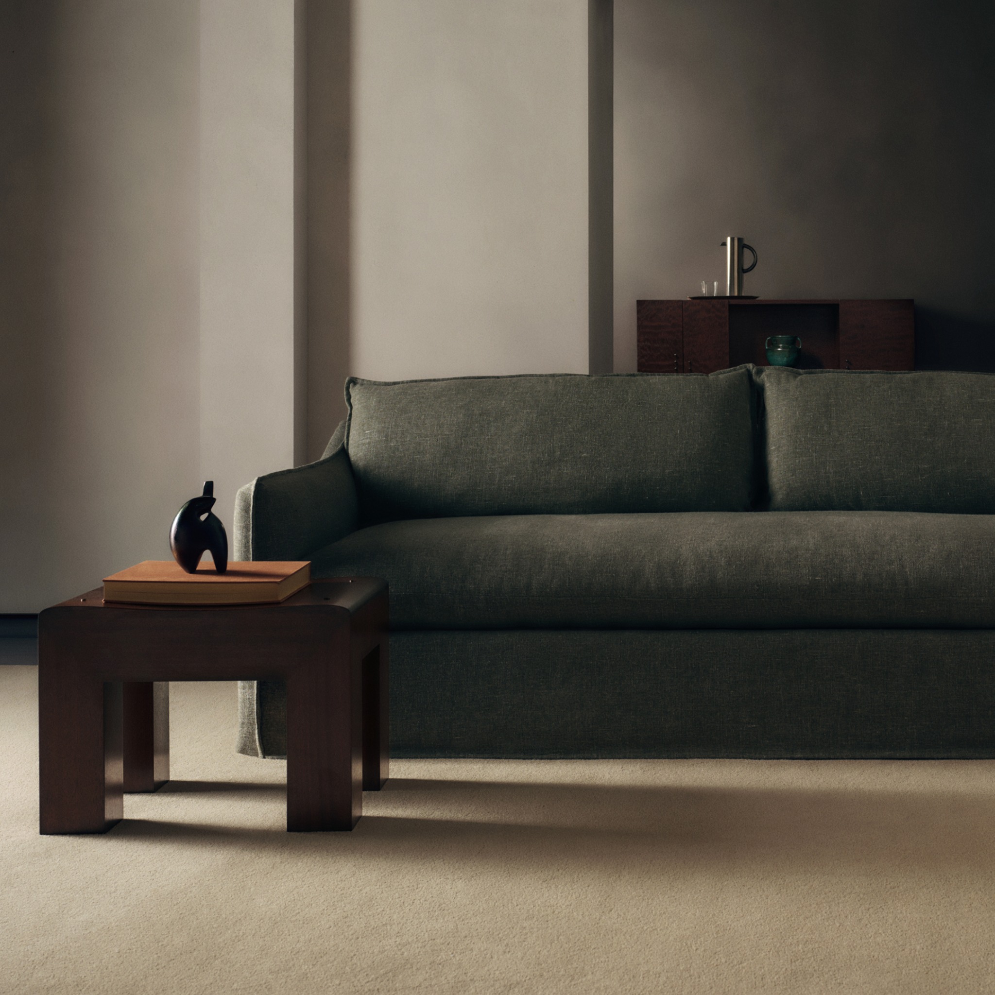 A dark, moody, and sophisticated living room scene is depicted. In the foreground, a dark wooden side table holds a stack of books topped with a small, abstract black sculpture. Behind the table, a large, deep green sofa with plush cushions dominates the frame. In the background, a dark wooden credenza holds a metallic thermos and two glasses, with a small teal vase placed on top. The overall aesthetic is minimalist and elegant, emphasizing the craftsmanship and design of the furniture.