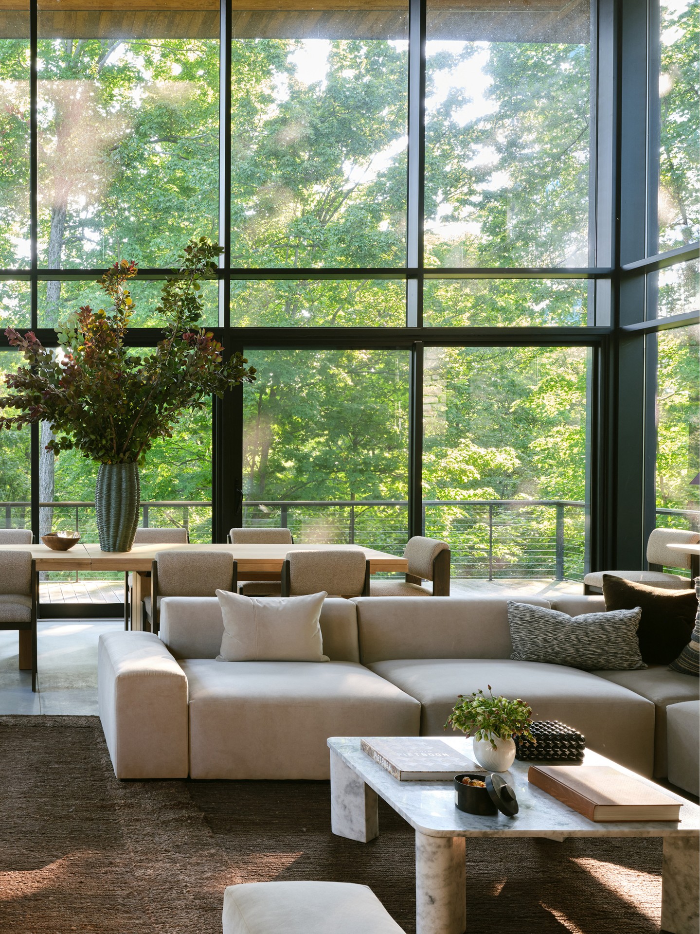 A wide shot of a modern living room with floor-to-ceiling windows overlooking a lush green forest. Sunlight streams through the trees and windows, illuminating a large, neutral-toned sectional sofa and a marble coffee table. The text overlay credits the interior design and photography for the Zionsville project, highlighting the Breuer Modular Sectional.