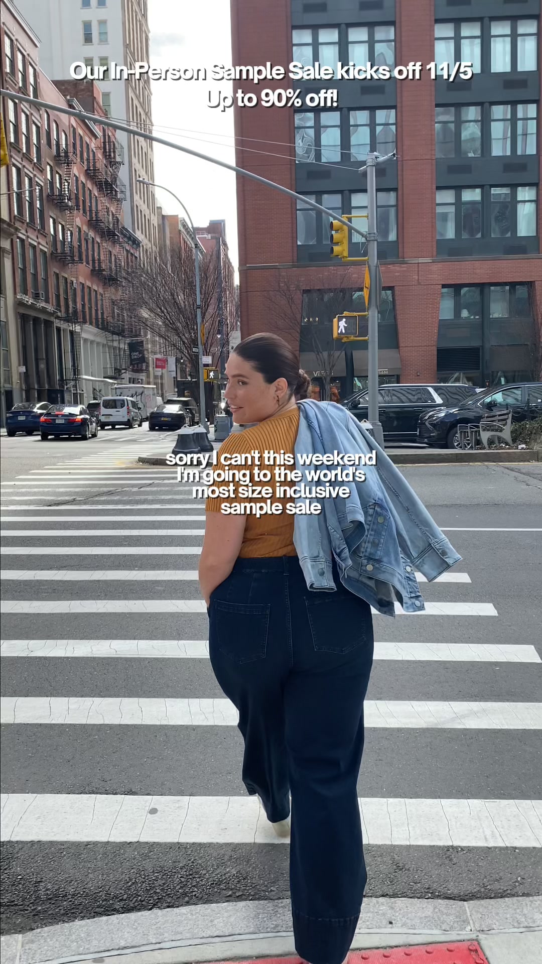 A woman walks across a city crosswalk, looking over her shoulder. She is wearing a mustard yellow top and dark wash jeans, with a denim jacket draped over her shoulders. White text overlays the top portion of the image, announcing "Our In-Person Sample Sale kicks off 11/5 Up to 90% off!". Below her, white text reads, "sorry I can't this weekender I'm going to the world's most size inclusive sample sale".