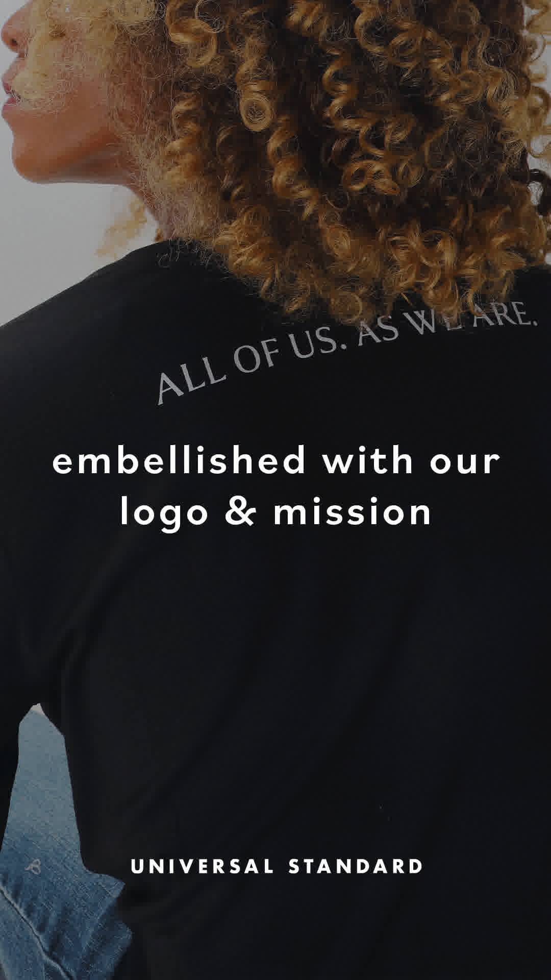The creative features a close-up of a person with curly blonde hair, wearing a black t-shirt. The t-shirt has white text on it that reads "ALL OF US. AS WE ARE." and below that, "embellished with our logo & mission". At the bottom of the image, the text "UNIVERSAL STANDARD" is displayed. The overall aesthetic is minimalist and focuses on the message and branding of the apparel.