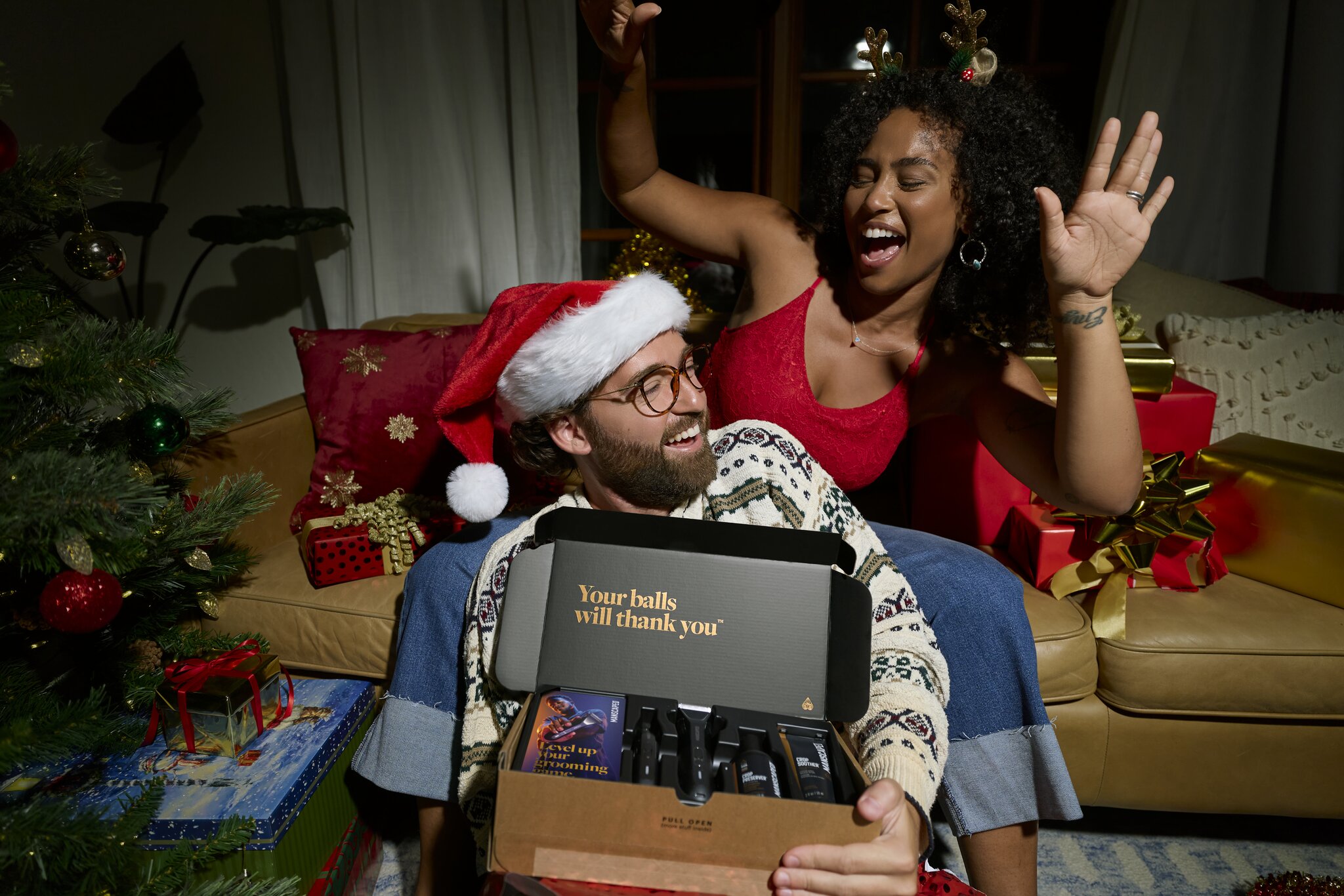 A man wearing a Santa hat and a festive sweater holds open a Manscaped grooming kit. A woman with reindeer antlers on her head is behind him, both smiling and looking excited. The inside of the box has the text "Your balls will thank you". The scene is decorated for Christmas with a tree and wrapped gifts, suggesting it's a holiday gift.
