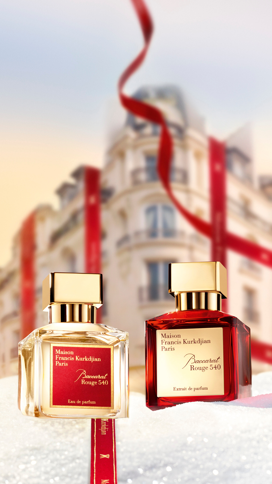 Two bottles of Maison Francis Kurkdjian Baccarat Rouge 540 perfume are displayed in a snowy setting, with a blurred Parisian building and red ribbons in the background. The left bottle is clear with a golden cap and a red label, while the right bottle is red with a golden cap and a cream-colored label. The text "Pour les fêtes, offrez l'inimitable." is overlaid on the image, suggesting a luxurious gift for the holiday season.