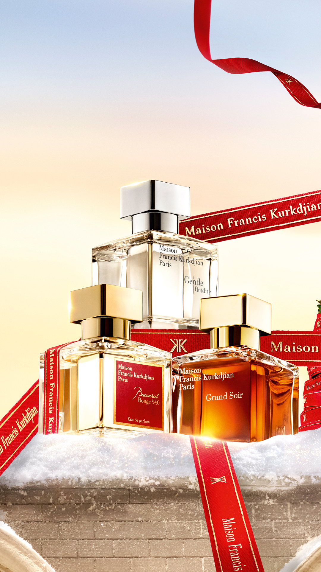 Three Maison Francis Kurkdjian perfume bottles are displayed on a snowy surface, adorned with red ribbons featuring the brand's logo. The background transitions from a warm, golden hue near the horizon to a soft blue sky. Text overlays on the ribbons read "Maison Francis Kurkdjian Paris" and "Gentle fluidity". The overall aesthetic evokes a festive, luxurious holiday atmosphere, suggesting a gift or seasonal offering.