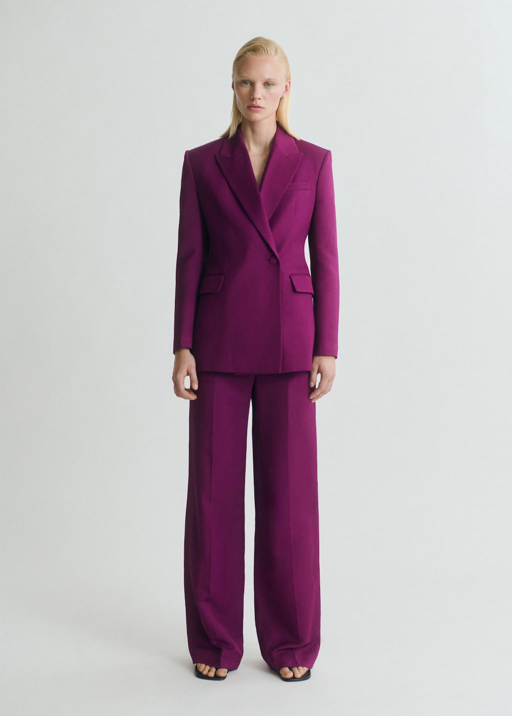 A full-body studio shot of a woman wearing a vibrant purple double-breasted blazer and matching wide-leg trousers. She stands against a plain white background, looking directly at the camera with a neutral expression. The text "¡Alerta, últimas unidades! Date prisa y hazte con nuestros productos más vendidos antes de que se agoten. 🚀" is overlaid on the image, emphasizing limited stock and encouraging a quick purchase.