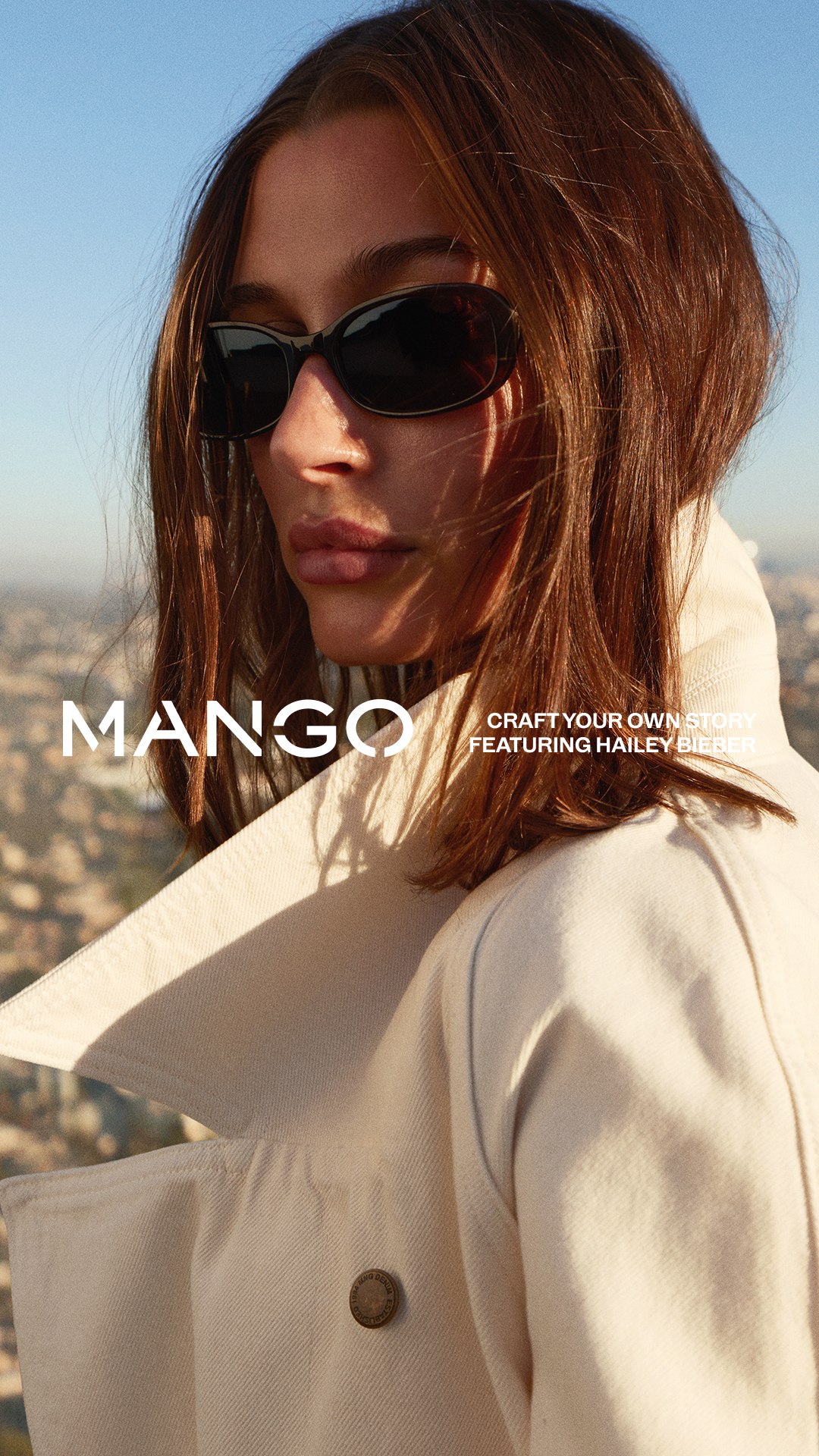 A close-up, low-angle shot of a woman wearing sunglasses and a cream-colored jacket. The background is a blurred cityscape under a clear blue sky. The Mango logo is prominently displayed on the left side of the image in white text. To the right of the logo, white text reads "CRAFT YOUR OWN STORY FEATURING HAILEY BIEBER." The overall aesthetic is chic and aspirational, suggesting a fashion campaign.