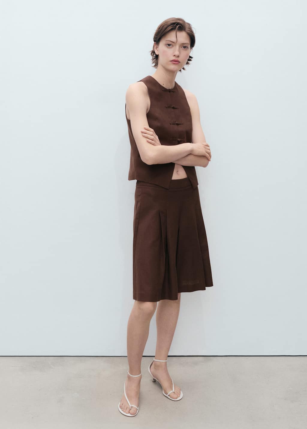 A female model stands against a plain white background, wearing a brown sleeveless top with frog closures and a matching pleated skirt. Her arms are crossed, and she looks directly at the camera. The text overlay asks, "What will you wear this spring?🌷" The ad promotes Mango's new collection for the spring season, suggesting light clothing, vibrant prints, and pastel colors as inspiration.