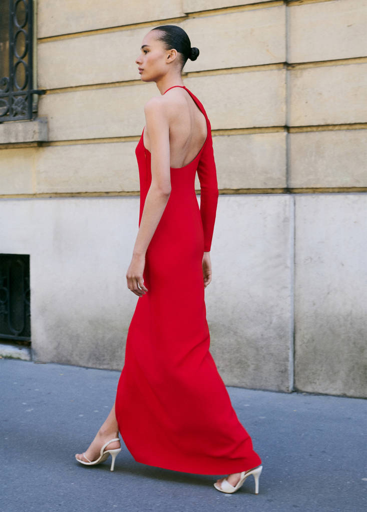 A woman walks from left to right, wearing a long, form-fitting red dress with an asymmetrical neckline and one long sleeve. She is wearing strappy white heels. The background is a textured, light-colored building facade. The text "Disfruta de esta selección de piezas en promoción. ¡Te están esperando en mango.com! 😉" is overlaid on the image.