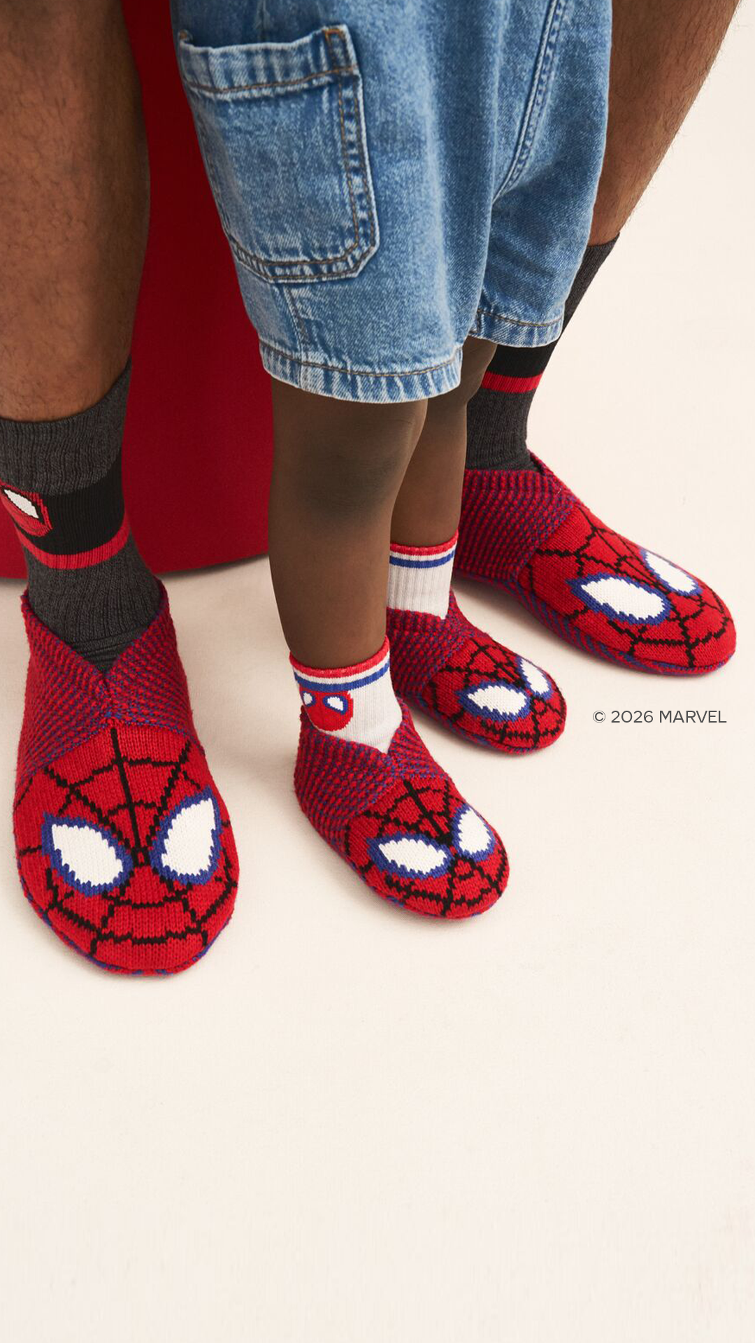 A close-up shot shows the lower legs and feet of an adult and a child, both wearing Spider-Man themed socks and slippers. The adult on the left wears dark grey socks with a Spider-Man mask design and red-and-black knitted slippers. The child in the center wears white socks with a Spider-Man mask and red-and-black knitted slippers. The background is a plain cream color with a hint of red on the left. The text "© 2026 MARVEL" is visible in the bottom right corner.
