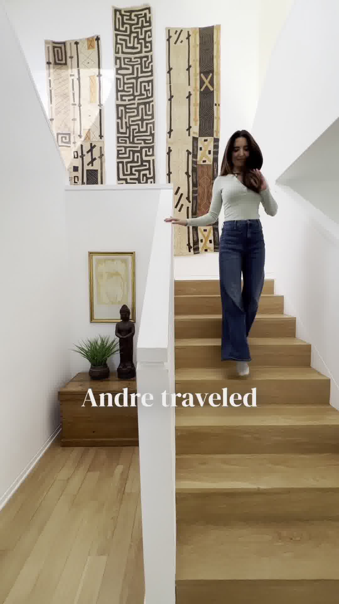 A woman in a light-colored long-sleeved shirt and dark wide-leg jeans walks down a wooden staircase in a modern home. She is smiling and looking towards the camera. The walls are white, and there are three large, decorative textile hangings with geometric patterns on the wall behind her. On the landing below the stairs, there is a wooden chest with a plant and a statue on it. White text overlaid on the bottom of the image reads "Andre traveled". The hook "You’re more powerful than you know. 🌟" is displayed prominently at the top of the creative.