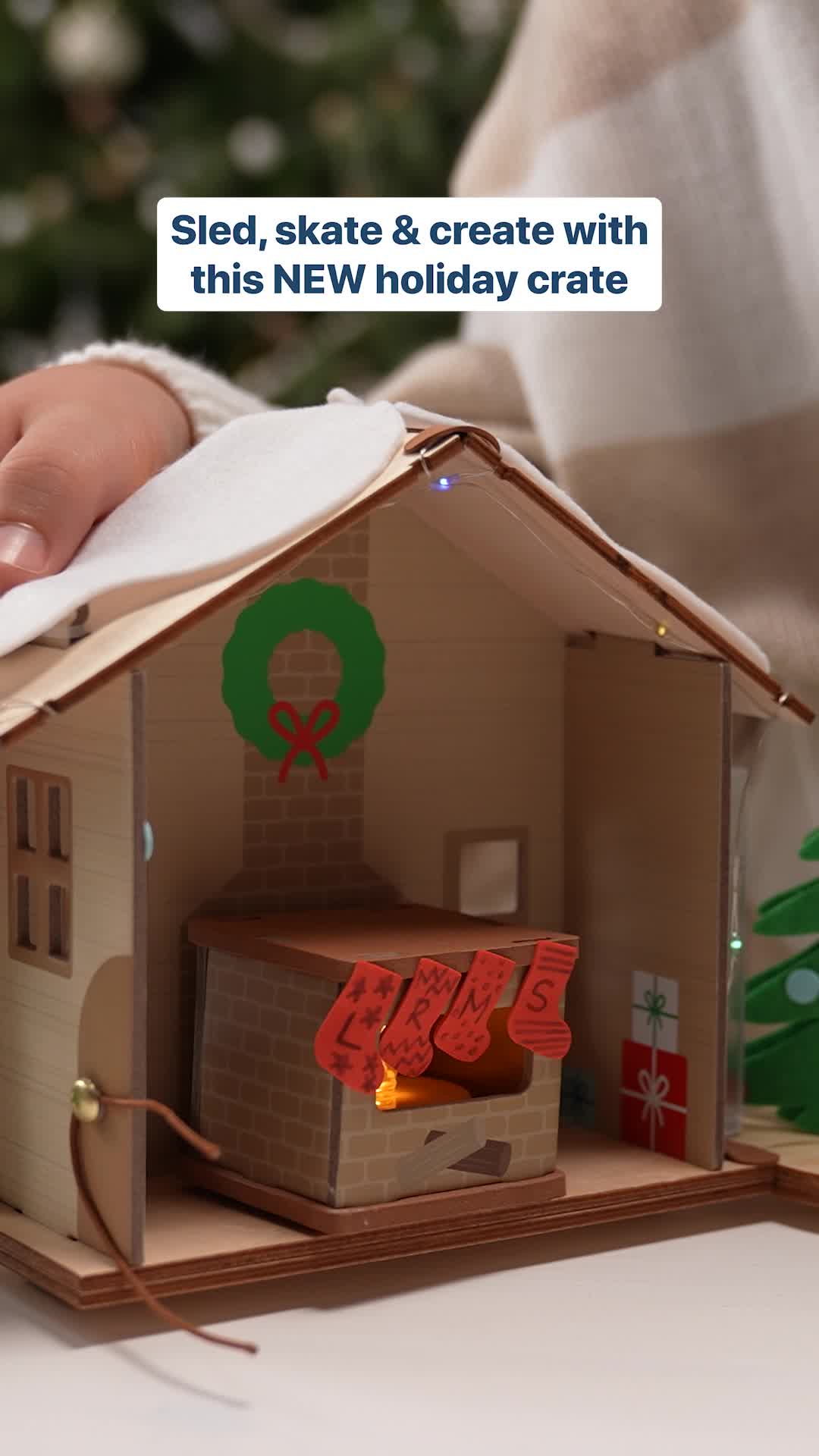 A close-up shot of a miniature wooden house decorated for the holidays. The house has a fireplace with stockings hanging, a wreath on the wall, and tiny Christmas lights. Text overlay reads "Sled, skate & create with this NEW holiday crate." The copy mentions KiwiCo Advent Calendars and encourages shopping before they sell out.