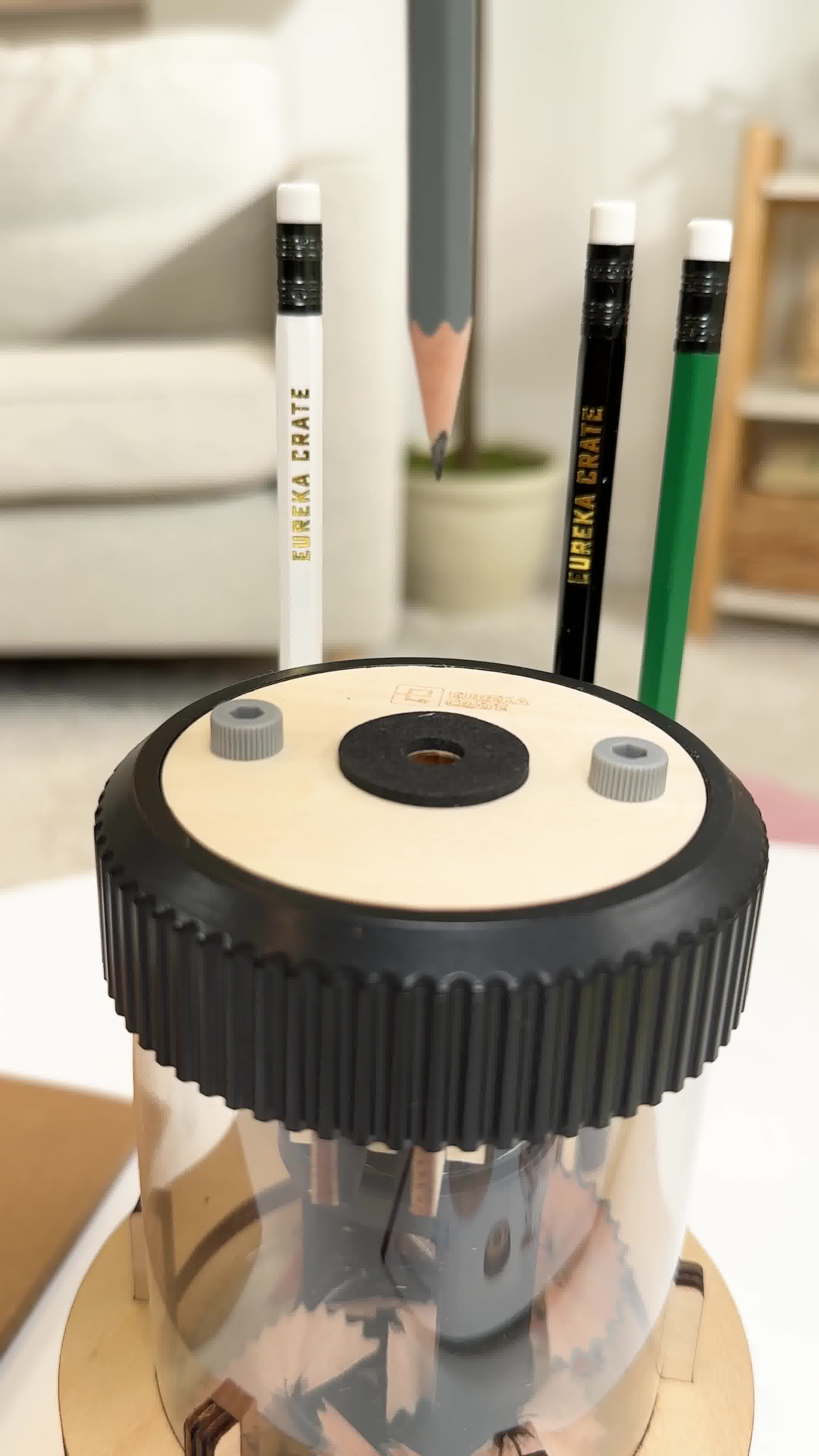 A close-up shot of a pencil sharpener with a clear plastic base showing pencil shavings. Several pencils, including one white, one black, and one green, are inserted into the top of the sharpener. The text "Like a regular pencil sharpener, but make it ⚡electric⚡(and DIY)." is overlaid on the image. The background is softly blurred, showing a hint of furniture and a plant.