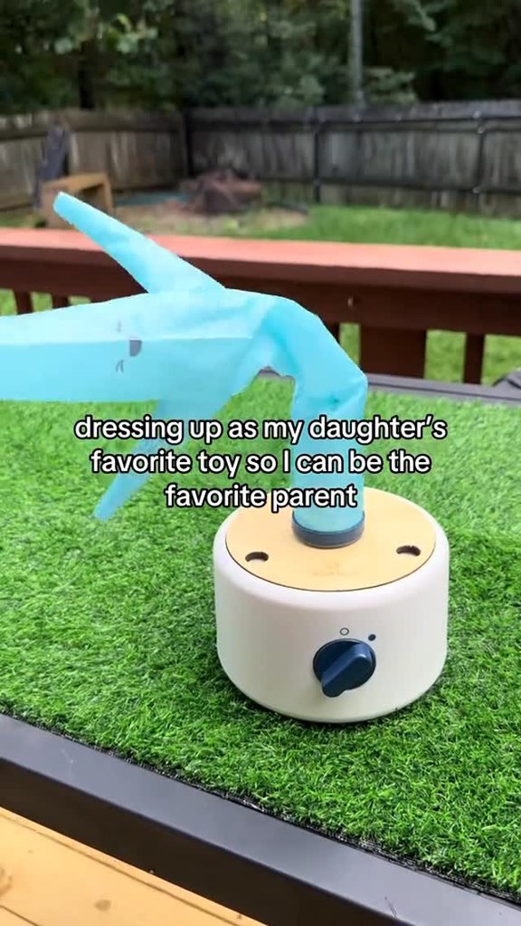 The image shows a close-up of a blue plastic toy resembling a dinosaur's head attached to a white base with a blue dial. The toy is positioned on a patch of artificial grass, with a wooden deck and a blurred backyard scene in the background. White text overlays the image, reading "dressing up as my daughter's favorite toy so I can be the favorite parent." The ad copy mentions being the favorite parent and offers a "Wind Play Lab" with a low stock alert.