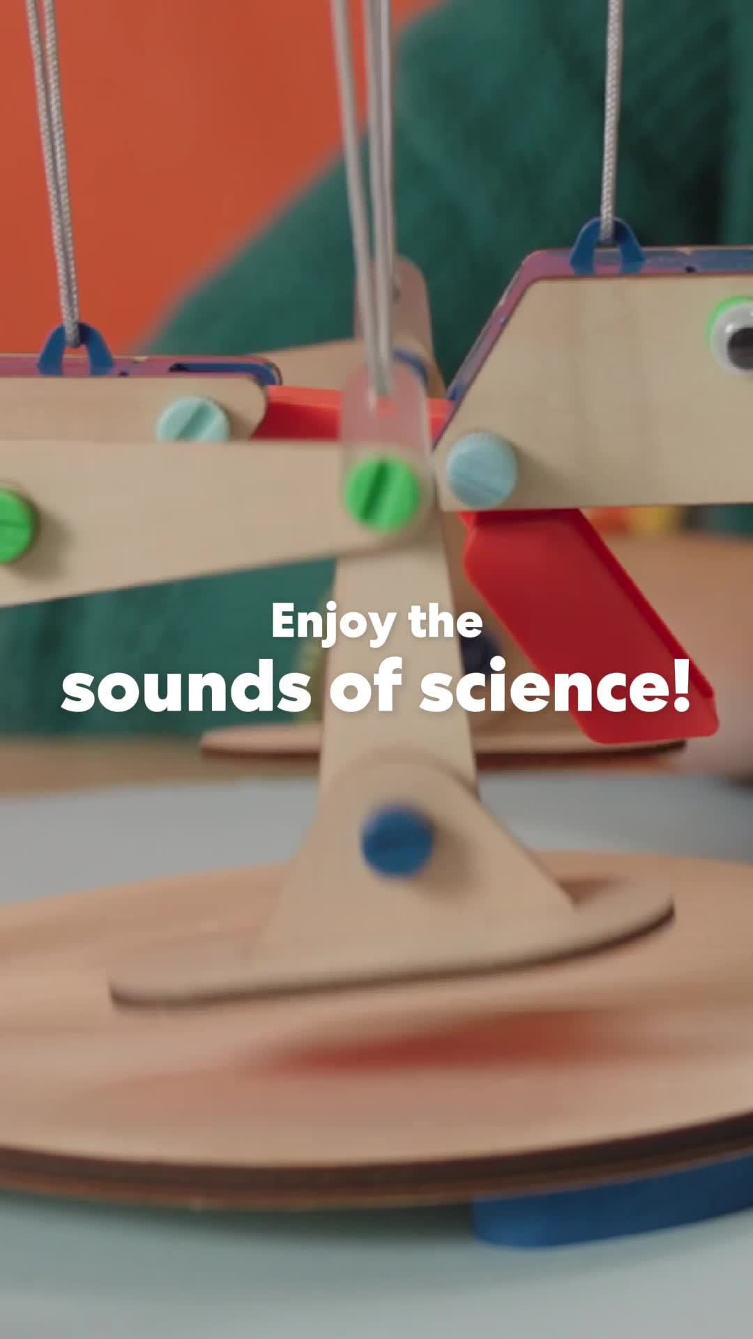 A close-up shot of a wooden science toy with colorful pegs and moving parts. White text overlays the image, reading "Enjoy the sounds of science!". The toy appears to be in motion, suggesting a demonstration of its function. The background is out of focus, with an orange wall and a person wearing a green sweater visible.