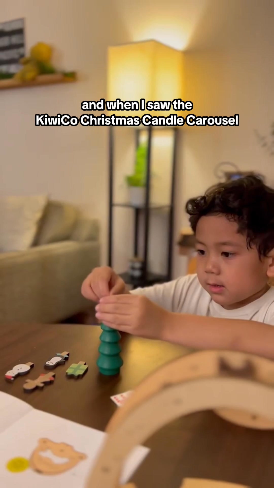 A young boy is shown playing with a Christmas-themed toy. The toy appears to be a candle carousel with small festive figures. On-screen text reads "and when I saw the KiwiCo Christmas Candle Carousel". The background shows a dimly lit room with a lamp and furniture, creating a cozy atmosphere.