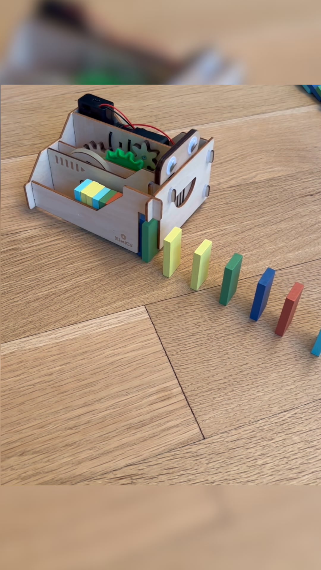 A wooden toy domino machine with googly eyes and a smiling mouth is shown on a wooden floor. The machine is loaded with colorful dominoes and is set up to push them onto a line of dominoes already on the floor. The text overlay reads "When Grandma finds the COOLEST gift 👀 Start Tinker Crate with the bestselling KiwiCo Domino Machine! 🎁".
