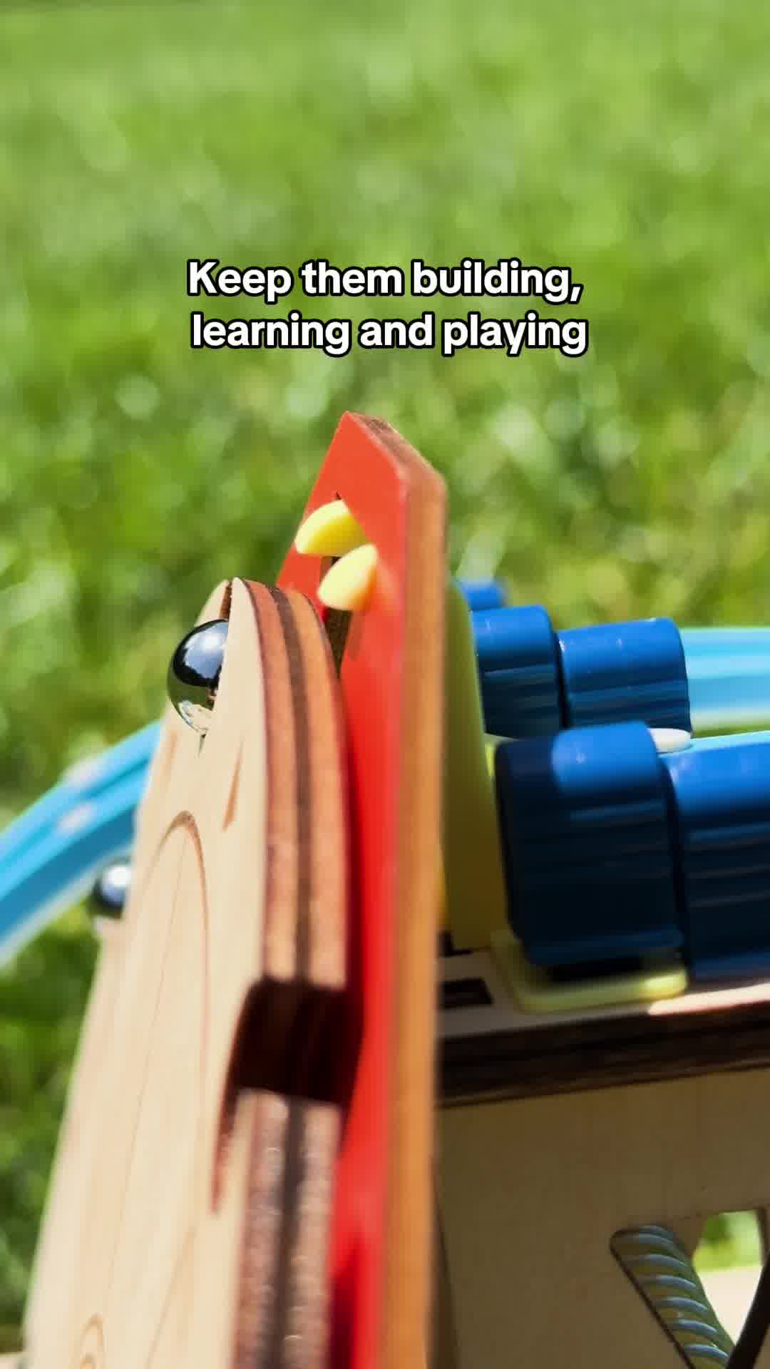 A close-up shot of a wooden toy with red and blue plastic components, possibly a marble run or construction set, is shown outdoors with a blurred green background. White text overlaid on the image reads "Keep them building, learning and playing." The focus is on the toy's construction and the text highlights its educational and entertainment value.