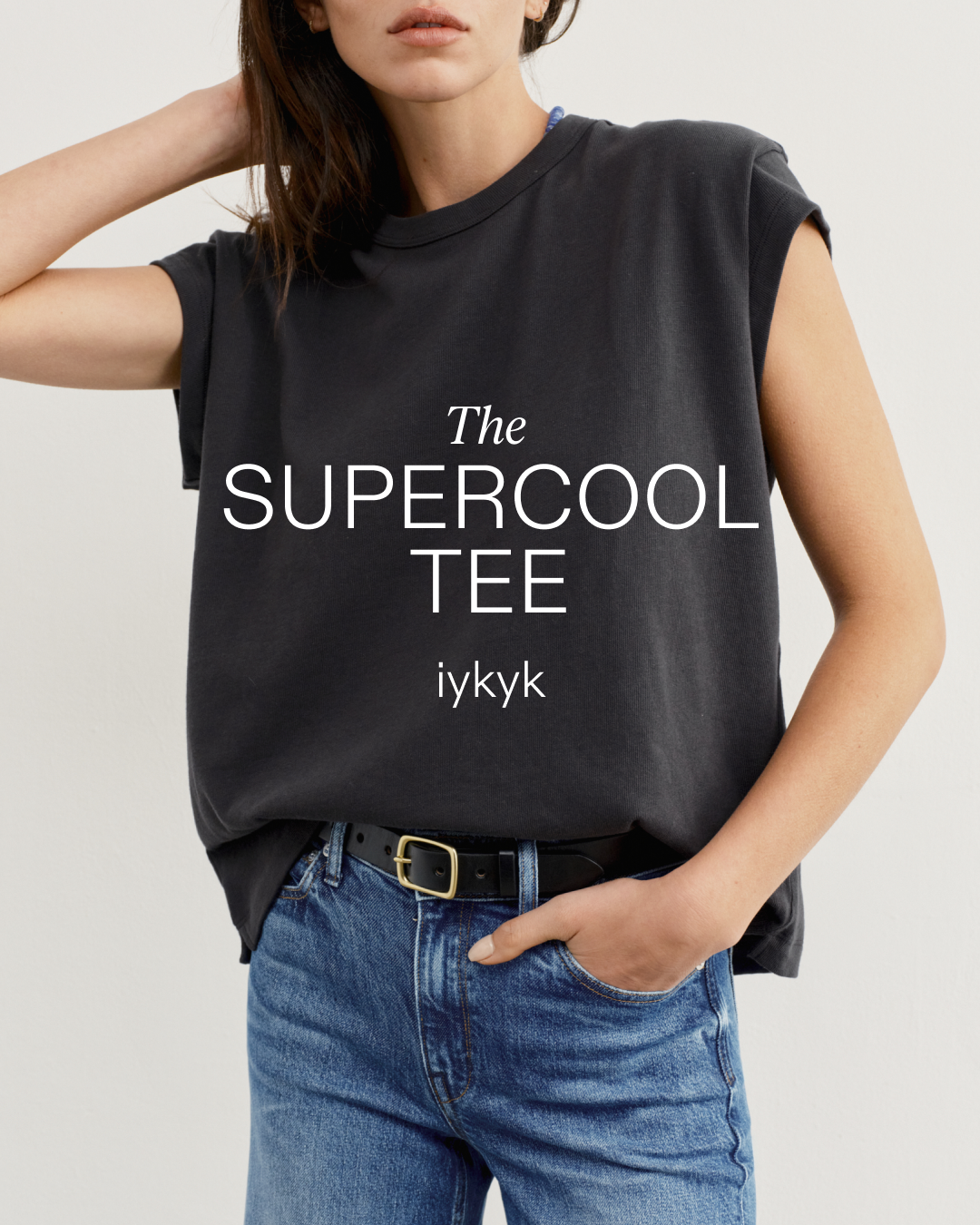 A woman is shown from the chest down, wearing a dark grey t-shirt and blue jeans. The t-shirt has the words "The SUPERCOOL TEE" printed in white, stylized font across the chest, with "iykyk" below it. The woman has her hand in her pocket and the other hand is touching her hair. The overall image is a clean, studio-style shot with a plain background.