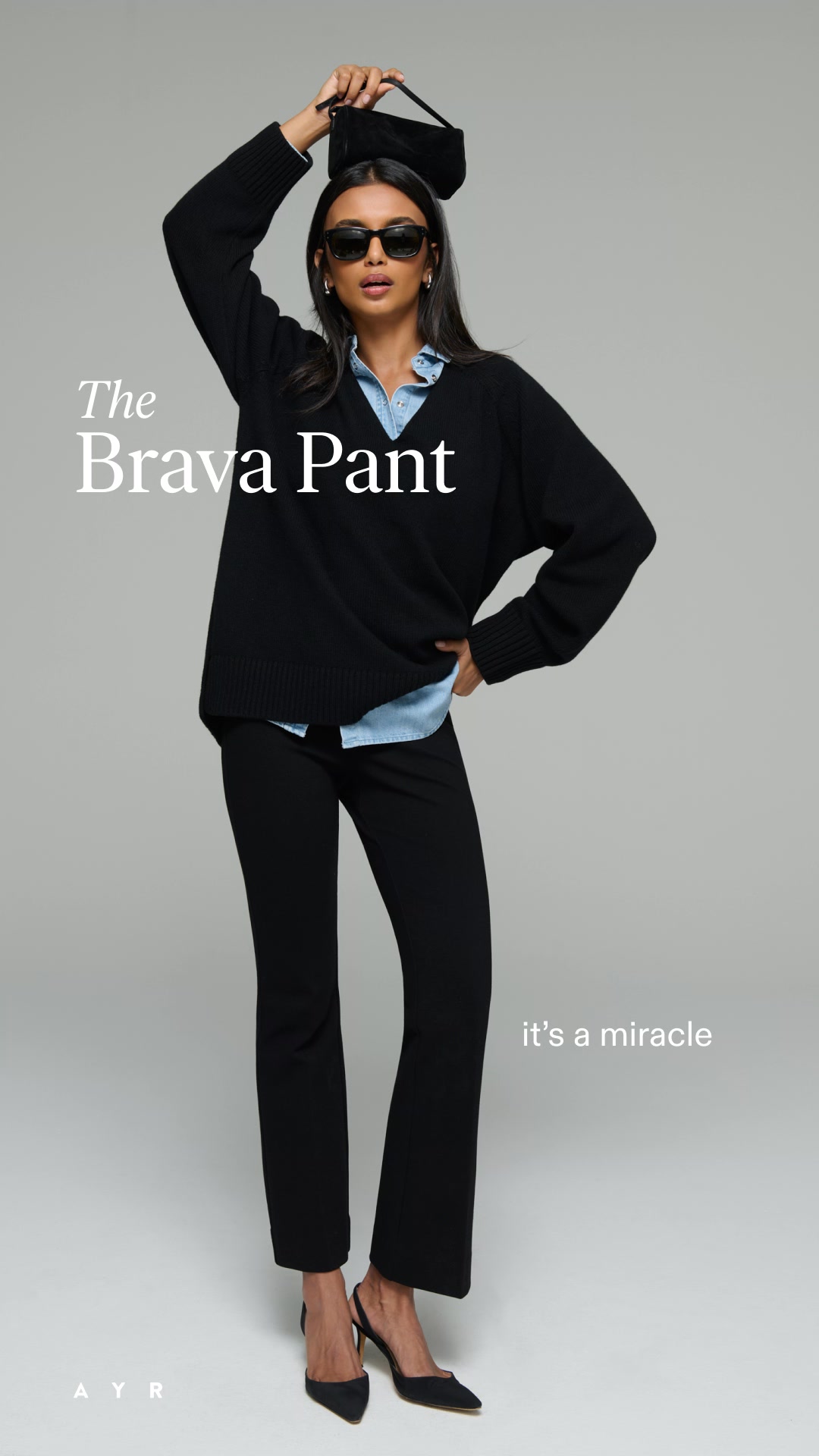 A woman stands against a plain grey background, posing with one hand on her hip and the other holding a small black purse above her head. She wears a black V-neck sweater, a light blue collared shirt underneath, and black flared pants. She also wears black sunglasses and black pointed-toe heels. White text on the left side of the image reads "The Brava Pant", and white text on the right side reads "it's a miracle". The brand logo "A Y R" is visible in the bottom left corner.