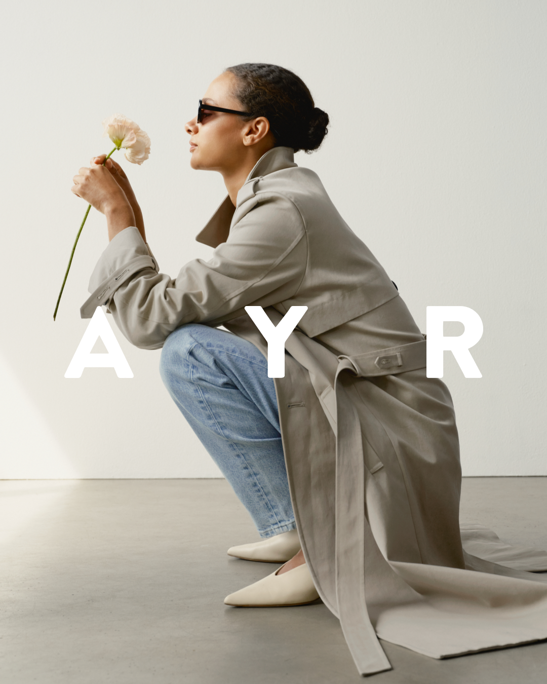 A woman wearing sunglasses and a trench coat crouches while holding a light pink flower up to her face. She is wearing light blue jeans and cream-colored pointed-toe flats. The letters "A Y R" are overlaid in large white font across the image. The background is a plain, light-colored wall and a grey floor. The overall aesthetic is minimalist and chic, suggesting a focus on timeless style and quality.