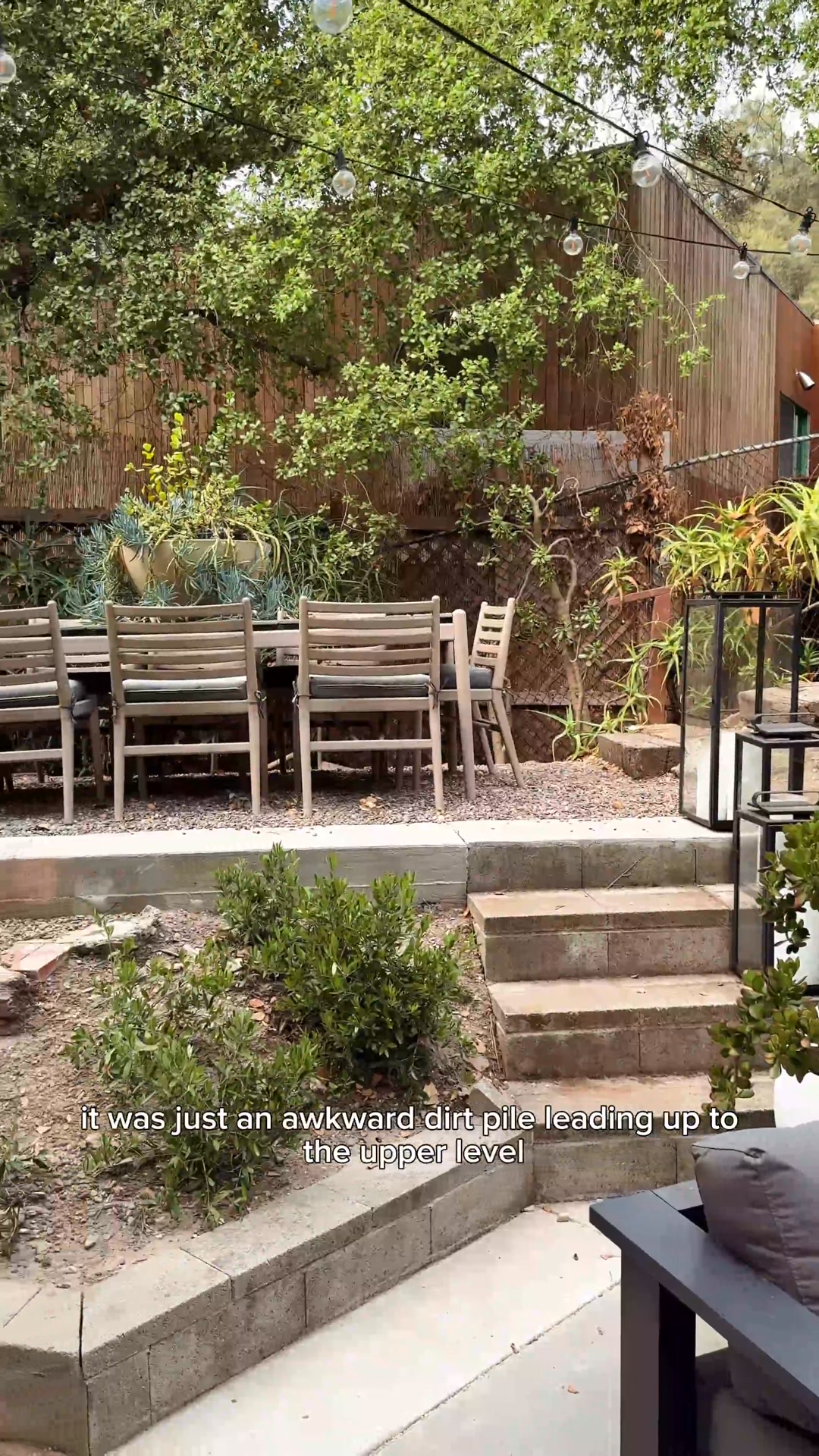 The image shows an outdoor patio area with a wooden dining table and chairs. In the foreground, there are stone steps leading up to a higher level, with greenery and landscaping. On-screen text reads, "it was just an awkward dirt pile leading up to the upper level," suggesting a home improvement project or transformation.