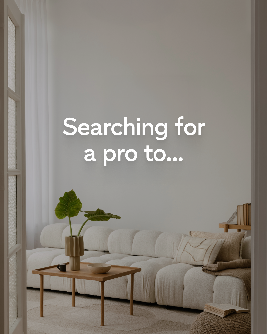 A minimalist living room with a plush white sofa and a wooden coffee table. The coffee table holds a vase with a large green leaf and a small black cup. White text overlaid on the image reads "Searching for a pro to...". The scene is bright and airy, with soft lighting and sheer curtains in the background.
